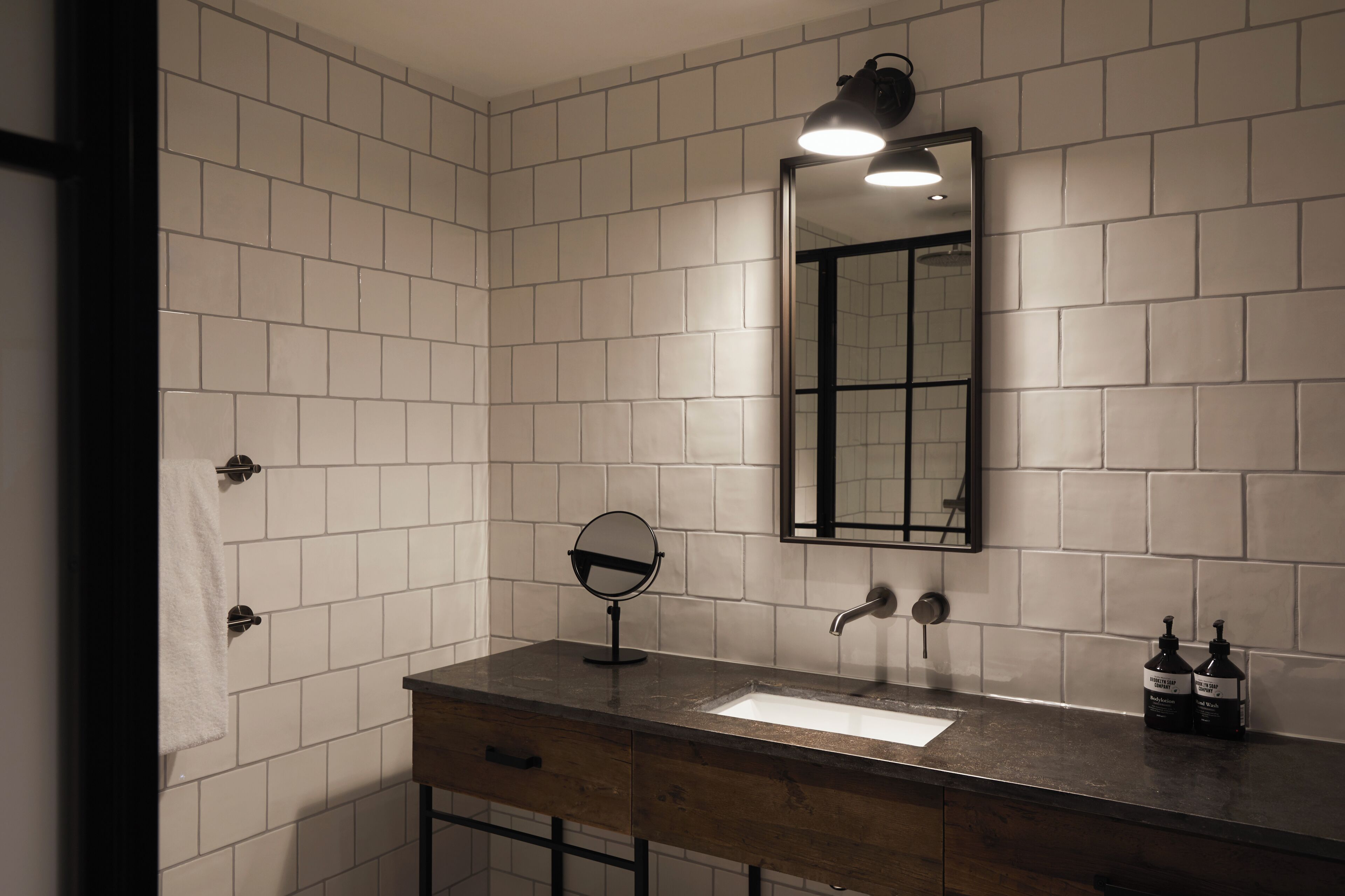 Un baño con paredes de azulejos blancos, un tocador de madera, un espejo, un lavabo, un grifo y dos botellas de jabón para manos.