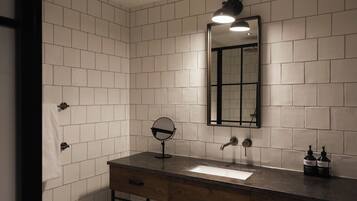 Un baño con paredes de azulejos blancos, un tocador de madera, un espejo, un lavabo, un grifo y dos botellas de jabón para manos.
