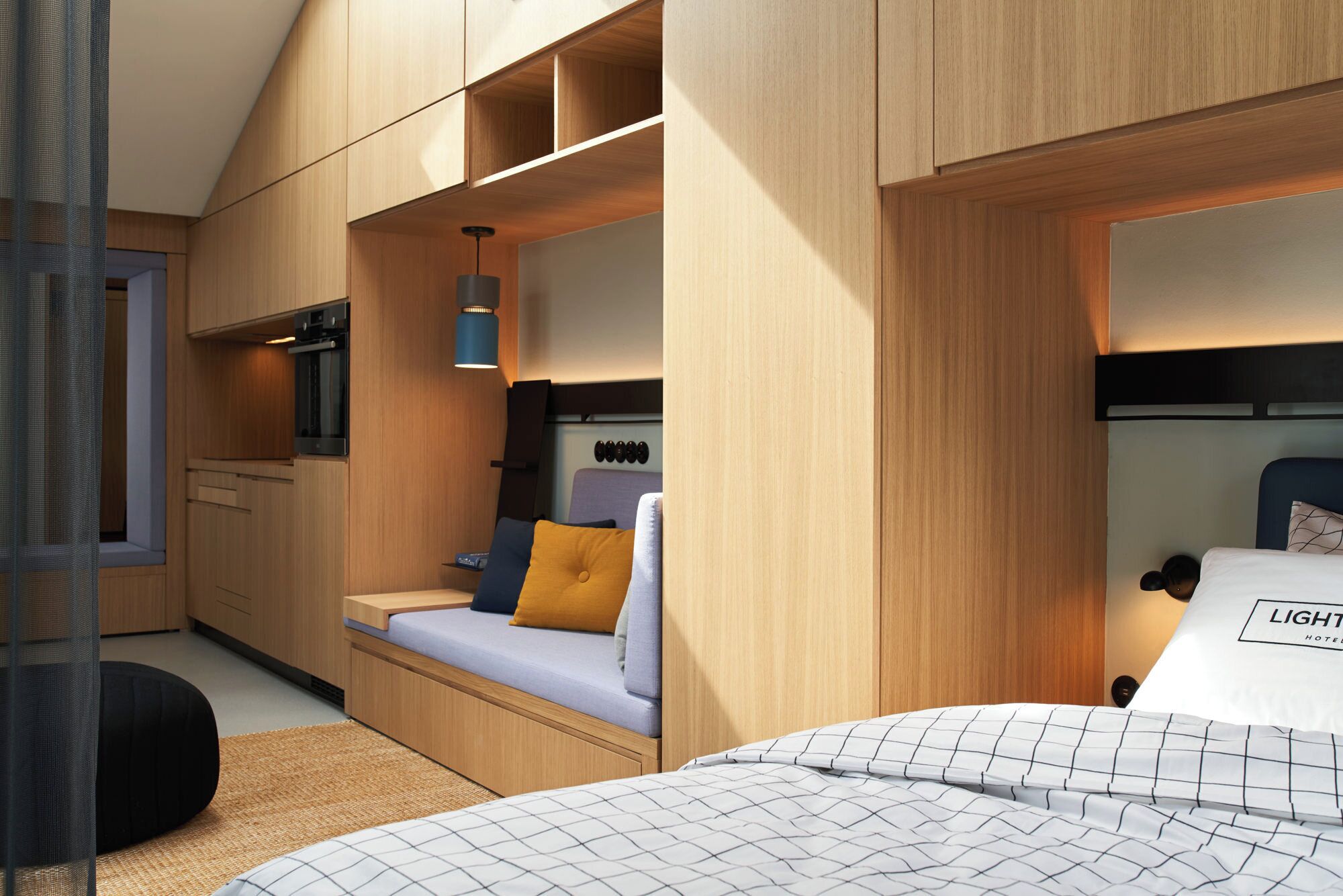A compact hotel room with a kitchenette, a bed with a patterned bedspread, and a small seating area with cushions.