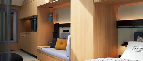 A compact hotel room with a kitchenette, a bed with a patterned bedspread, and a small seating area with cushions.