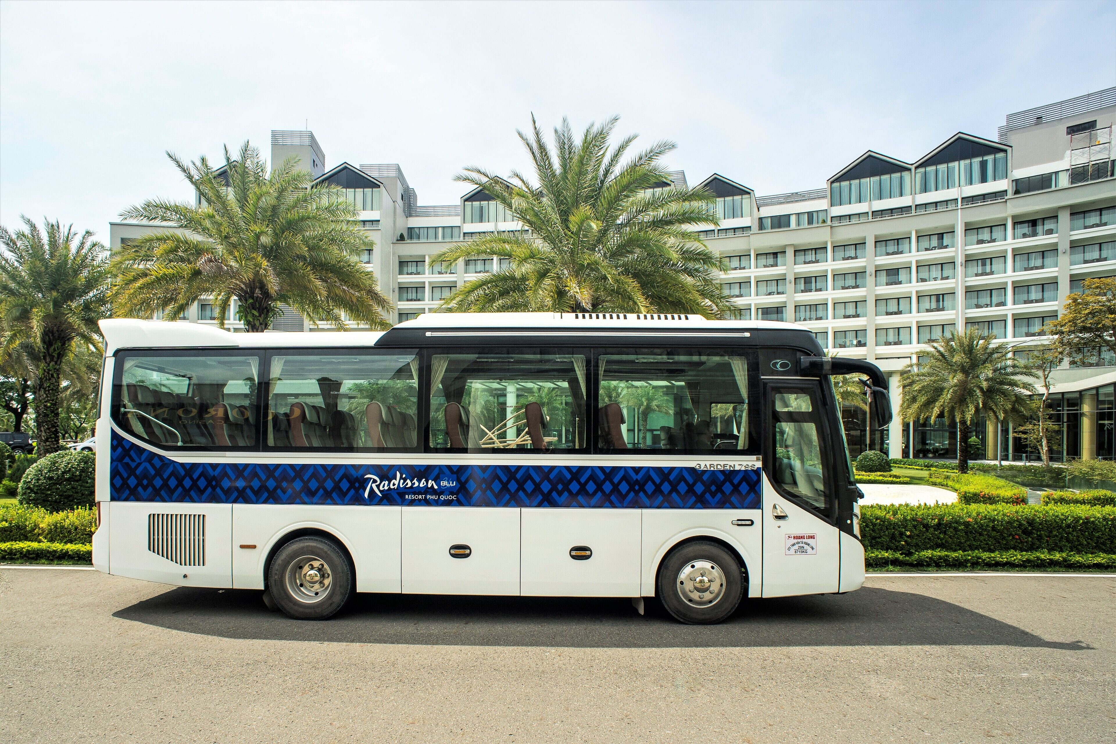A white bus with blue diamond patterns and the Radisson logo parked in front of a modern building with palm trees.
