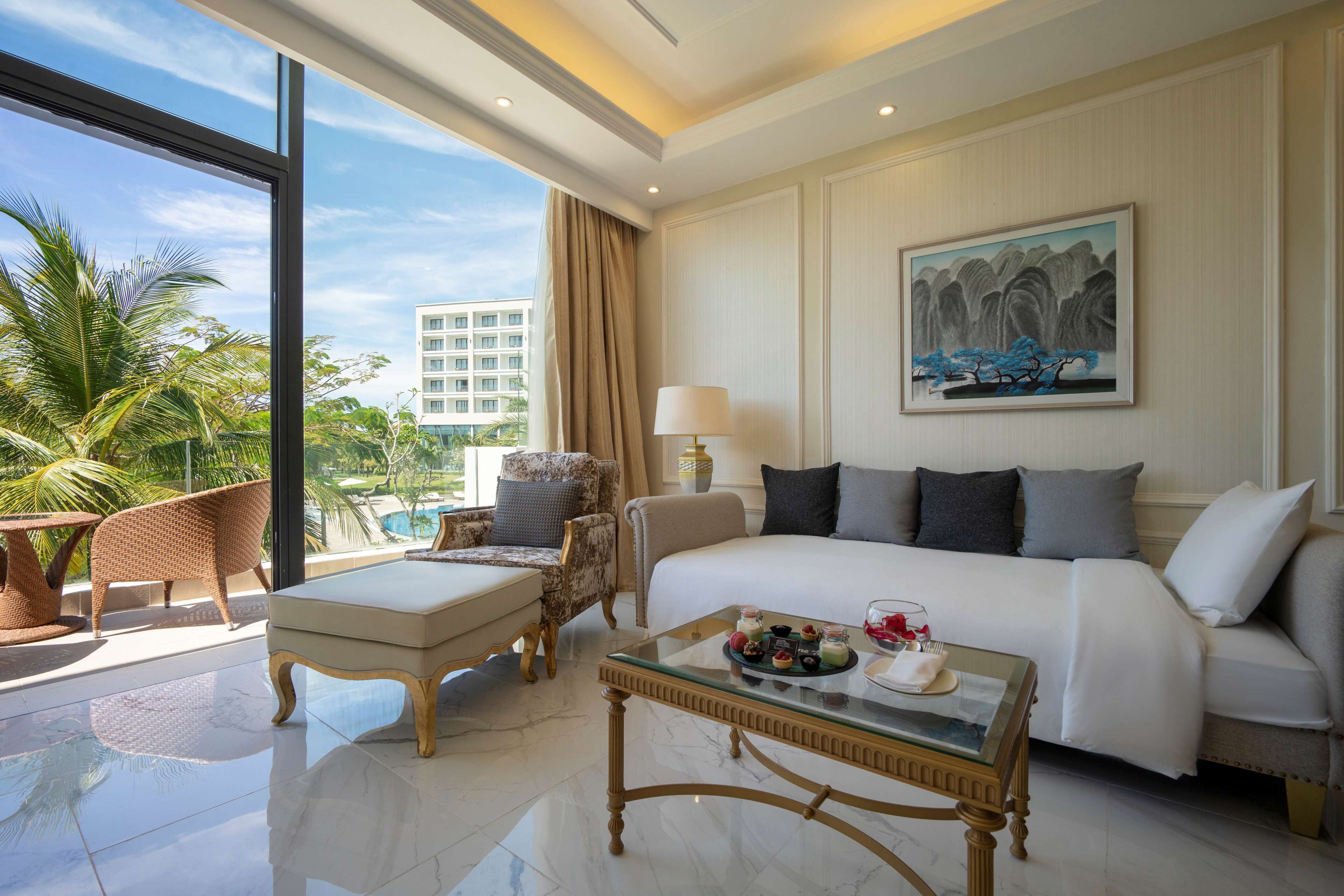 A hotel room with a bed, a glass coffee table, a sofa, and a chair. There is a painting on the wall and a view of palm trees and buildings outside the window.