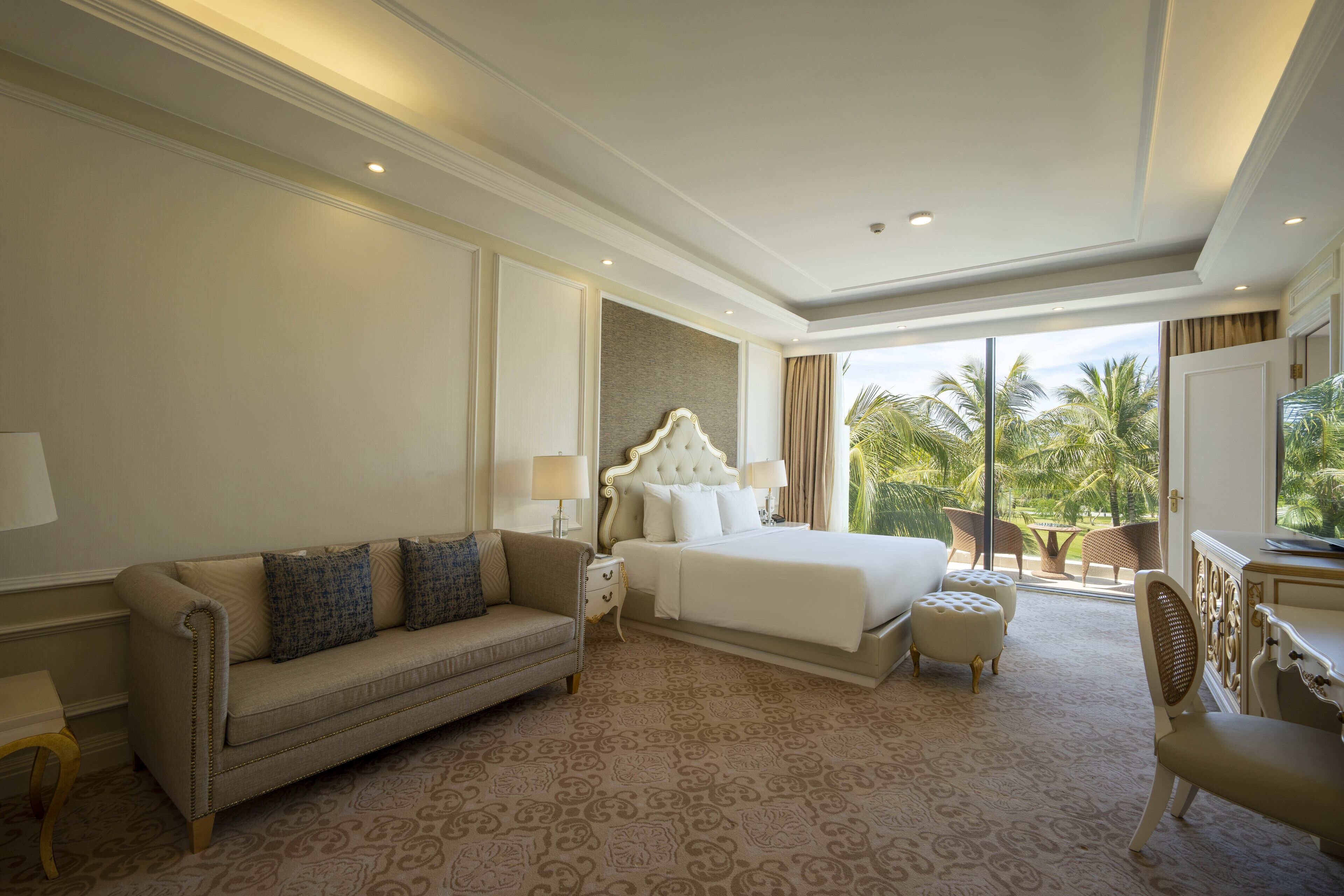 Habitación de hotel con una cama grande, un sofá, un escritorio y un balcón con vistas a palmeras.