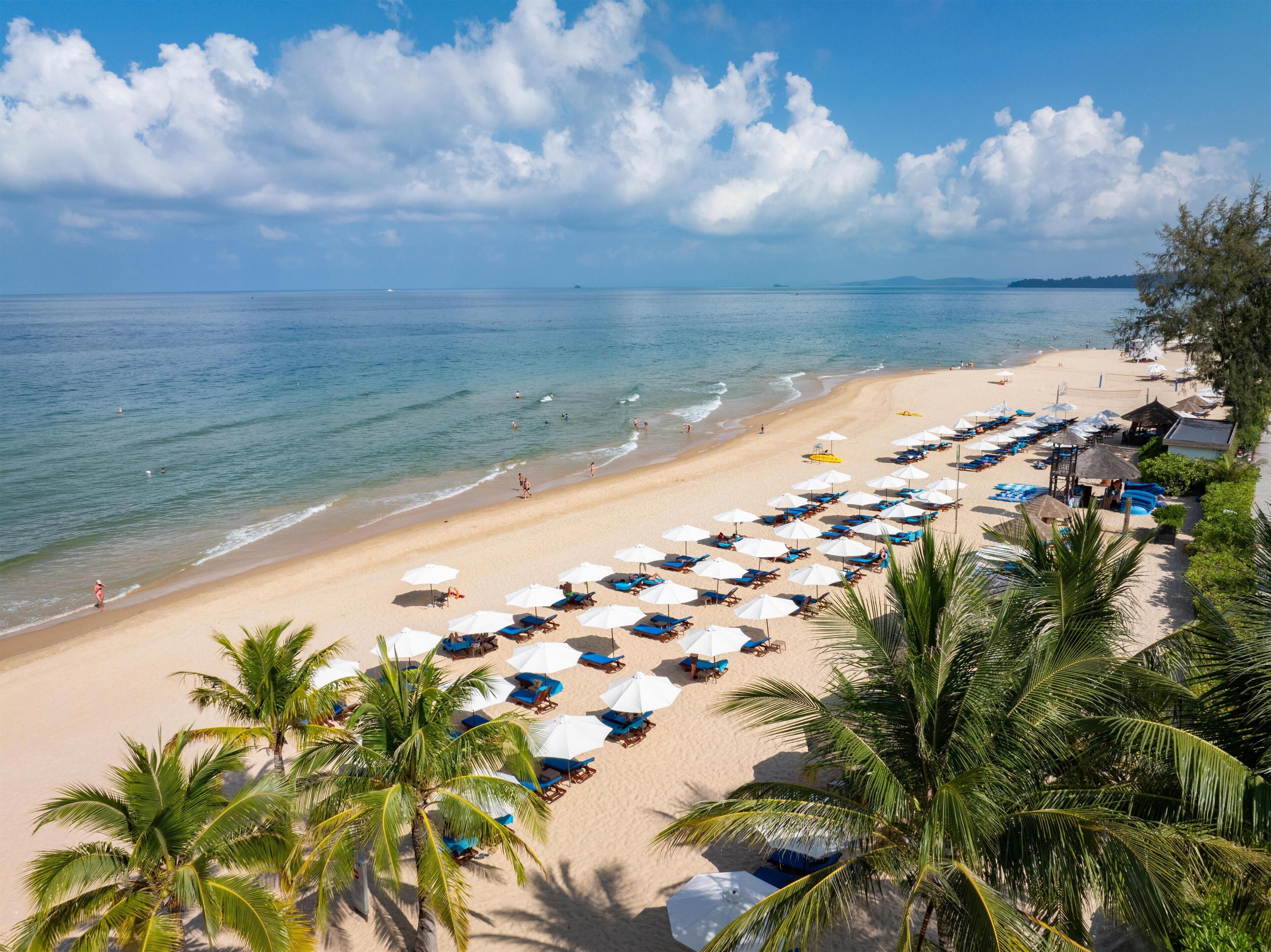 Een strand met rijen ligbedden en parasols, palmbomen en een helderblauwe lucht.