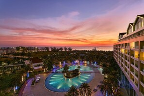 Piscina de un complejo turístico con diseño circular, rodeada de palmeras y sillas de descanso, con vistas a un cuerpo de agua al atardecer.