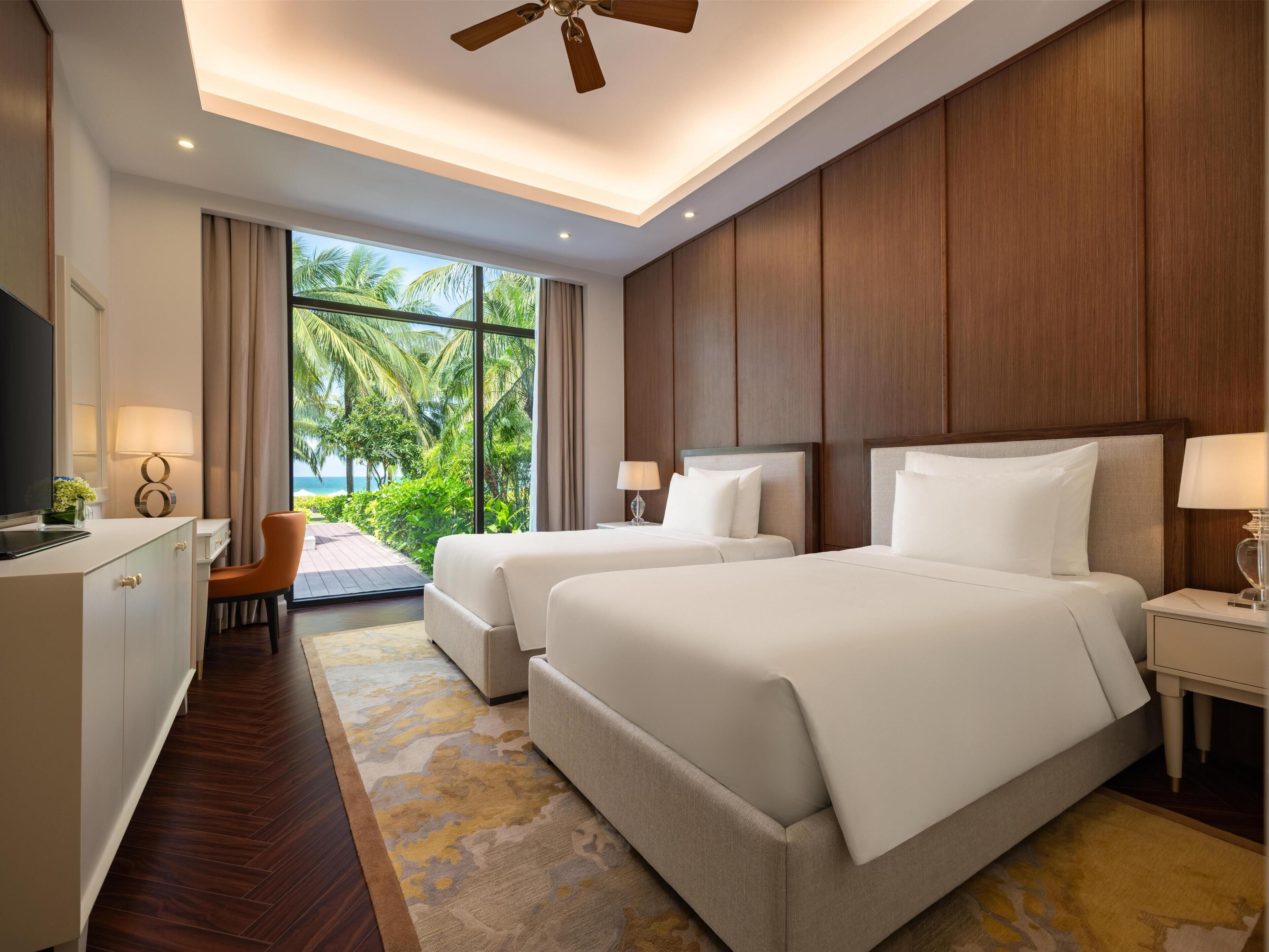 A hotel room with two beds, a desk, a chair, a TV, and a view of the outdoors.