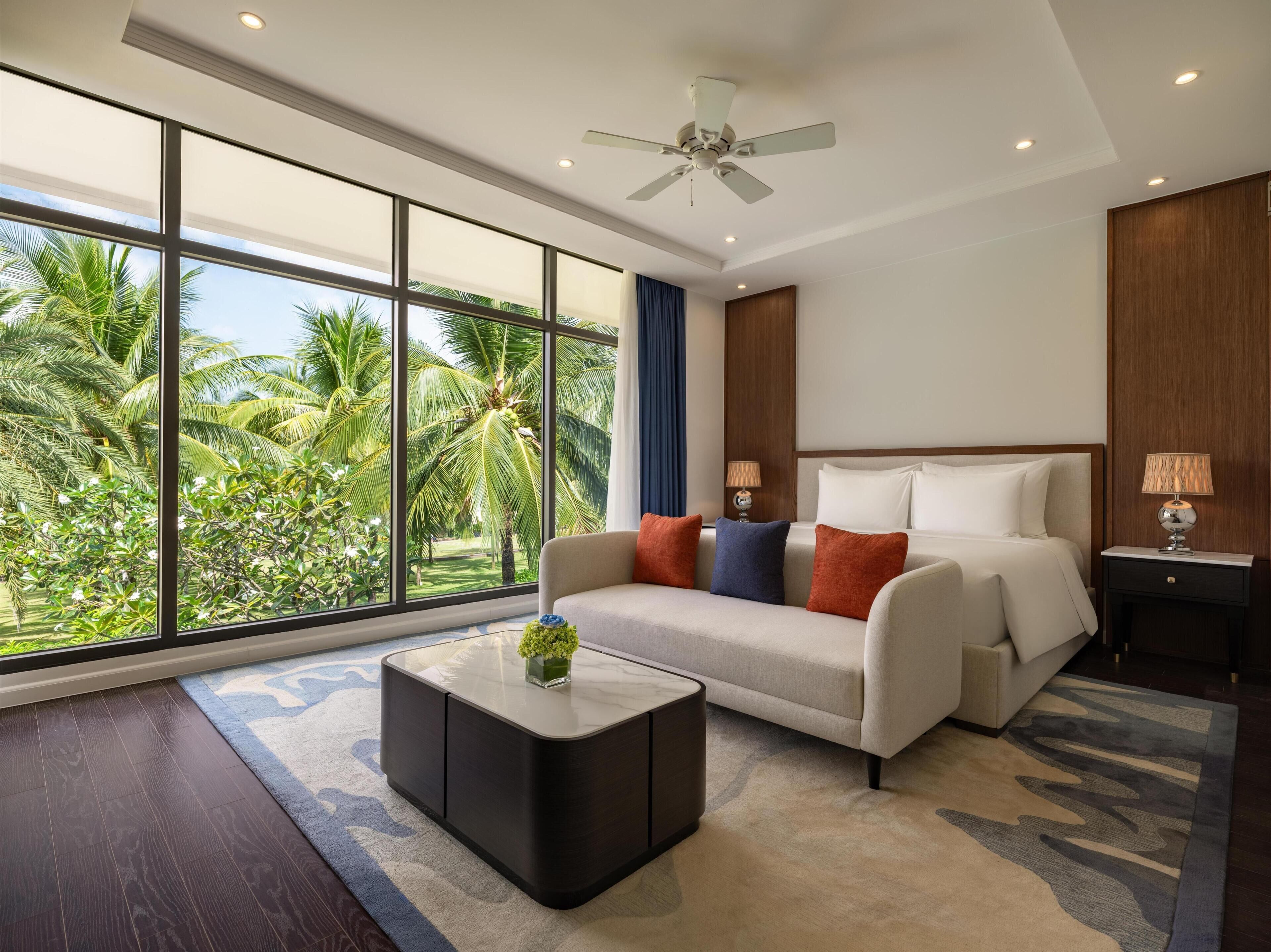 A modern hotel room with a large window, a ceiling fan, a bed, a sofa, a coffee table, and a nightstand.