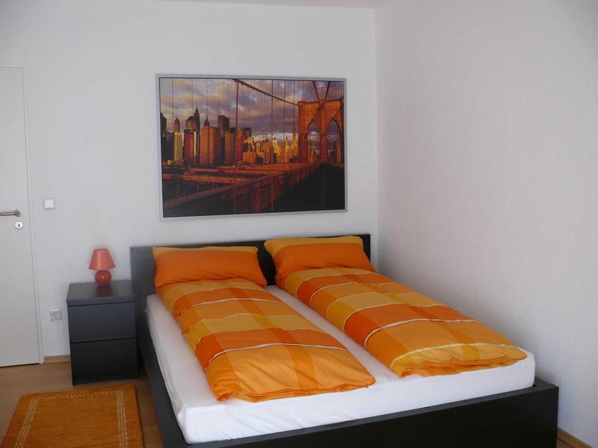 A bedroom with a bed, a nightstand, a lamp, and a cityscape painting on the wall.