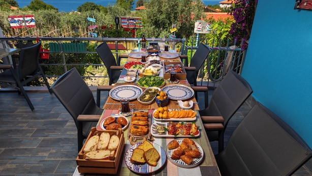 Une table dressée pour un repas, avec différents plats, entourée de chaises, et offrant une vue panoramique sur la mer et les collines.