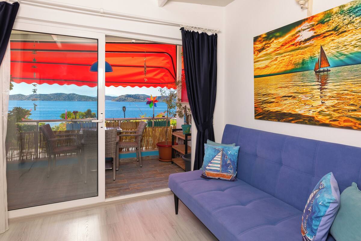 Een balkon met uitzicht op zee, een bank met kussens, en een schilderij van een zeilboot.