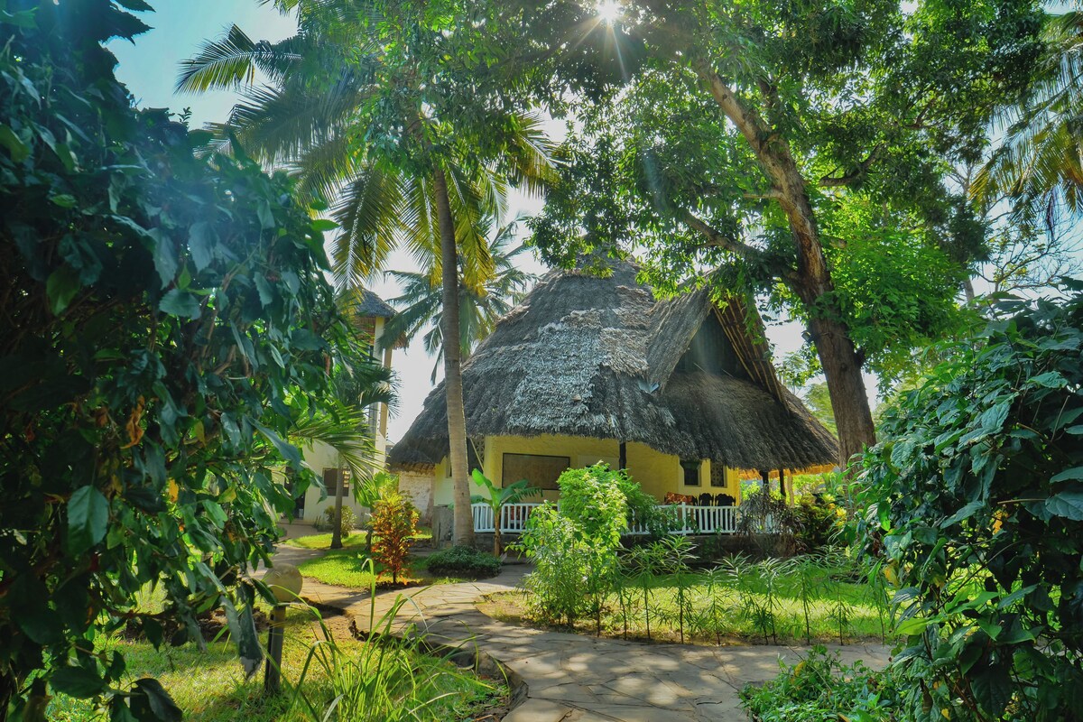 A thatched-roof house surrounded by lush greenery and a paved path.