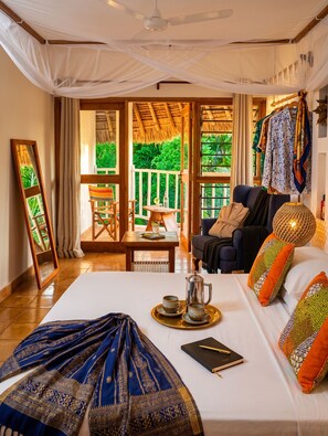 A spacious room with a large bed, a seating area with a table, and a view of lush greenery through large windows. - Upani in Daini (Diani Beach)