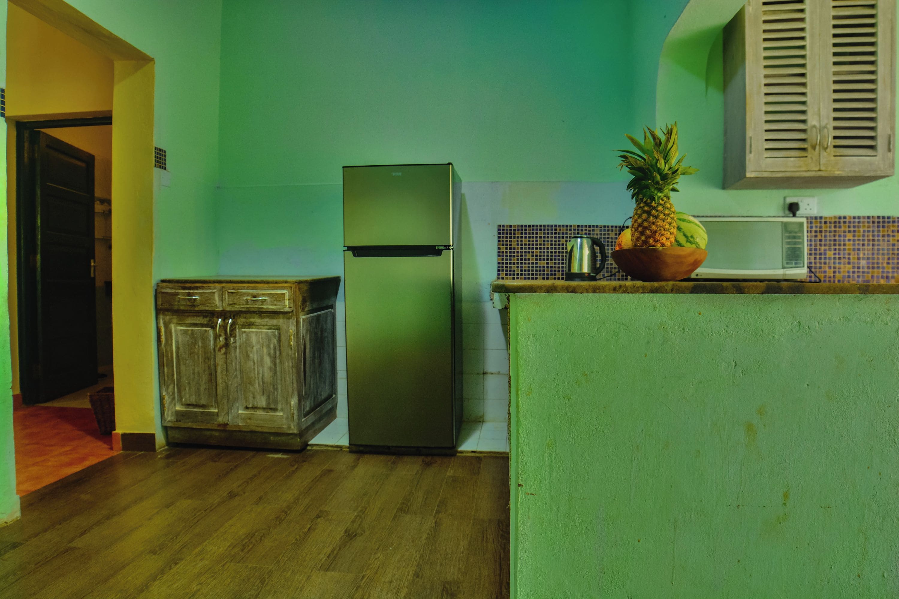 Une cuisine équipée d’un réfrigérateur, de meubles de rangement en bois, d’un plan de travail avec un revêtement en carreaux et d’un panier de fruits posé sur le comptoir.