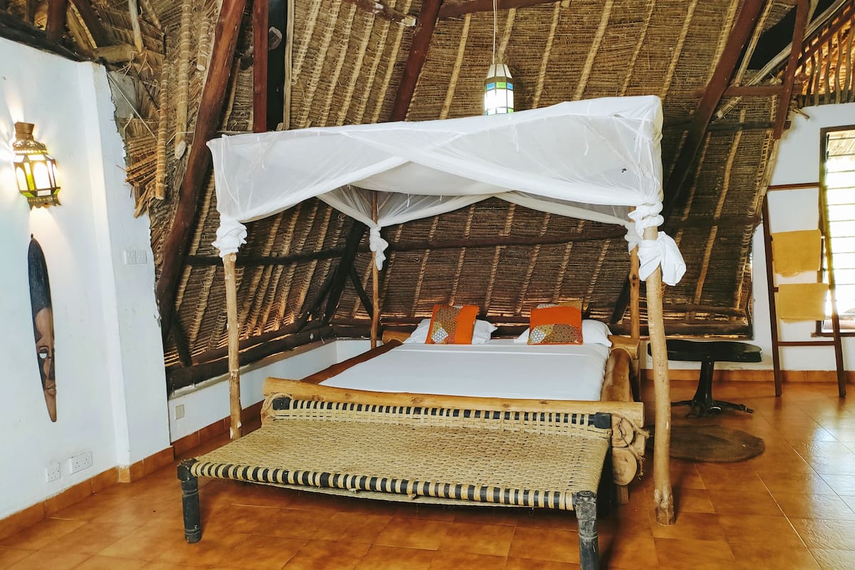 Quarto com telhado de palha, cama com dossel, um banco de fibra trançada e luminárias fixadas na parede.