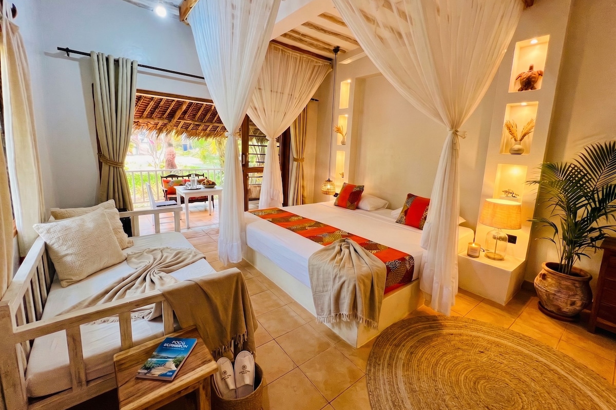 A bedroom with a bed, a sitting area with a chair and table, and a view of the outside.