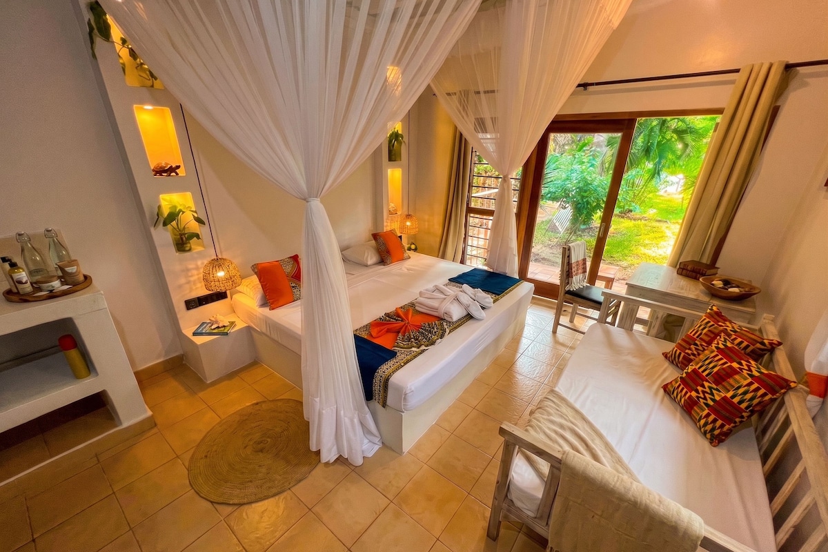 A room with a bed, a chair, a small table, and a window with a view of greenery.