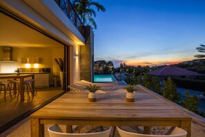 Una terraza de madera con piscina, mobiliario exterior y vistas al atardecer.