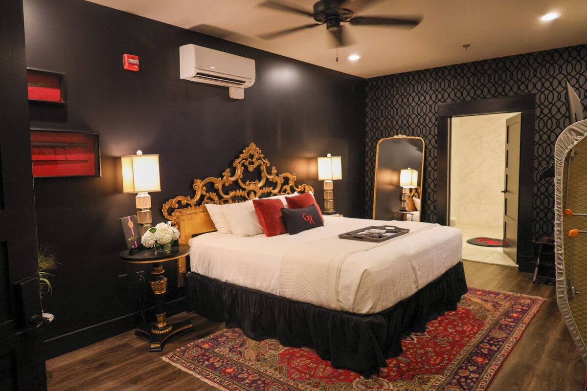 A hotel room with a large bed, ornate headboard, two bedside lamps, a red and black rug, and a ceiling fan.
