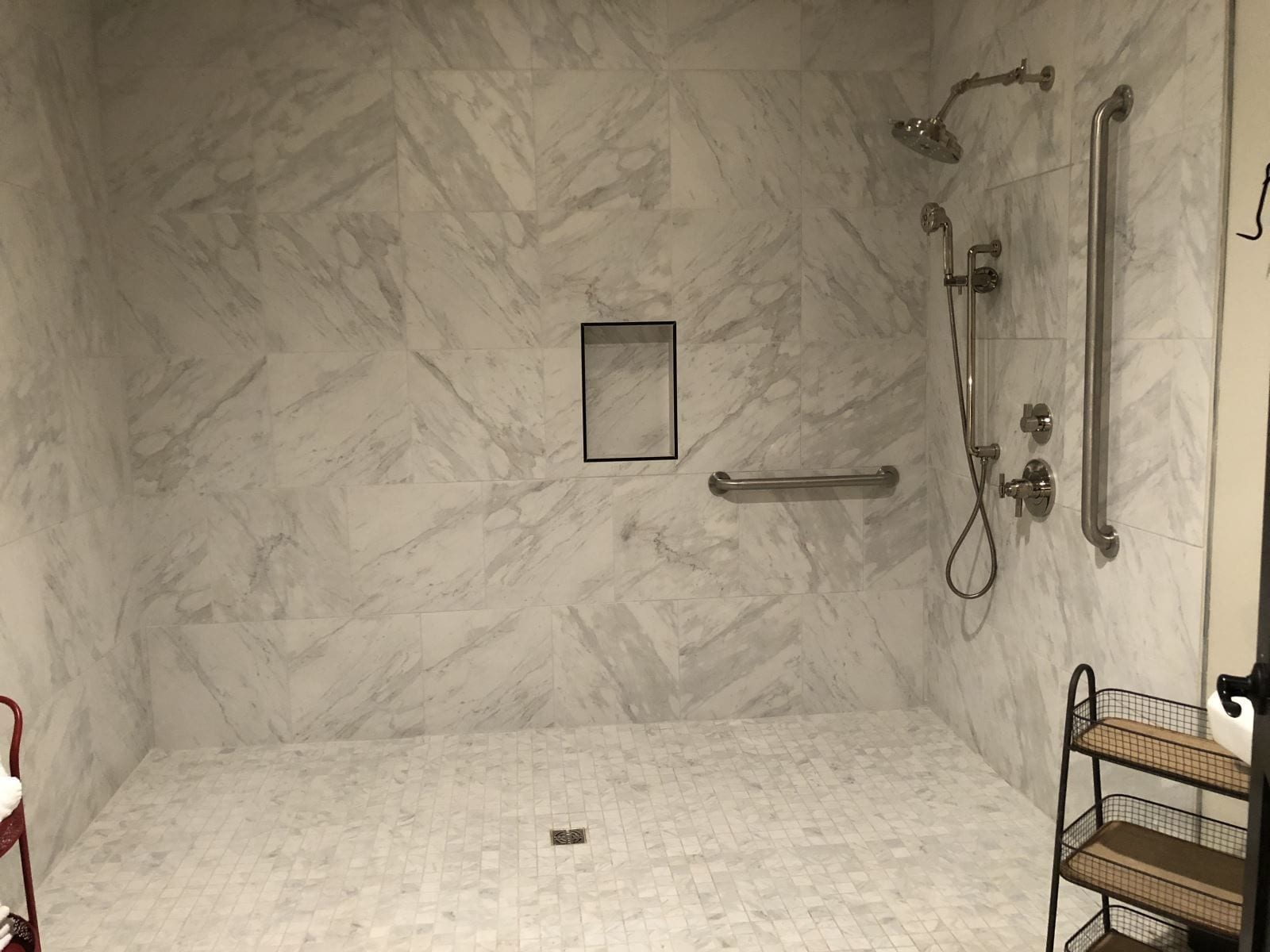 A bathroom with a marble wall, a shower with grab bars, and a small mirror.