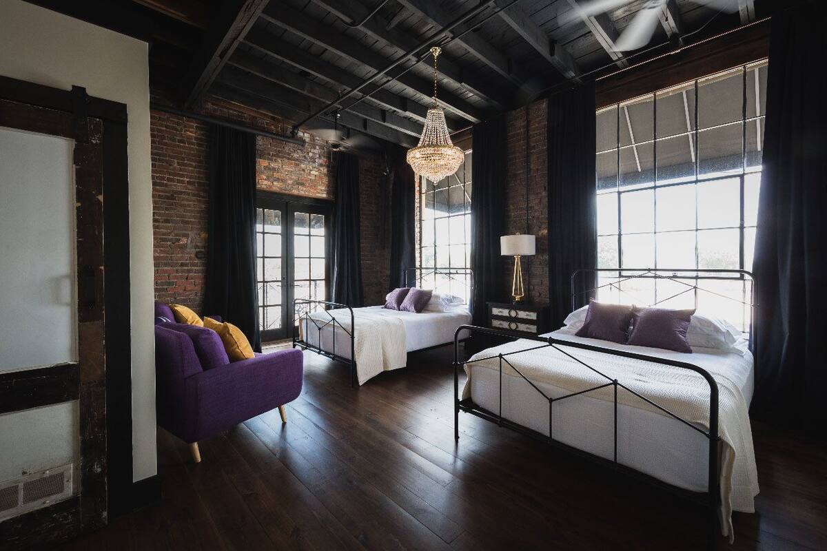 A modern room with two beds, a purple armchair, a chandelier, and large windows.