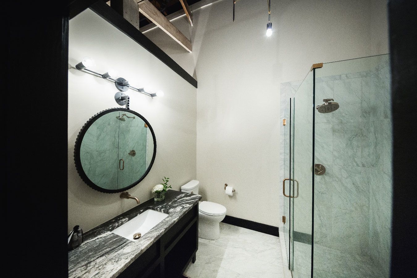 A modern bathroom with a round mirror, a marble sink, a toilet, and a glass shower enclosure.