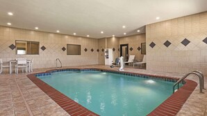 Una piscina interior con suelo de baldosas, borde de ladrillo rojo y una zona de estar con sillas y una mesa.
