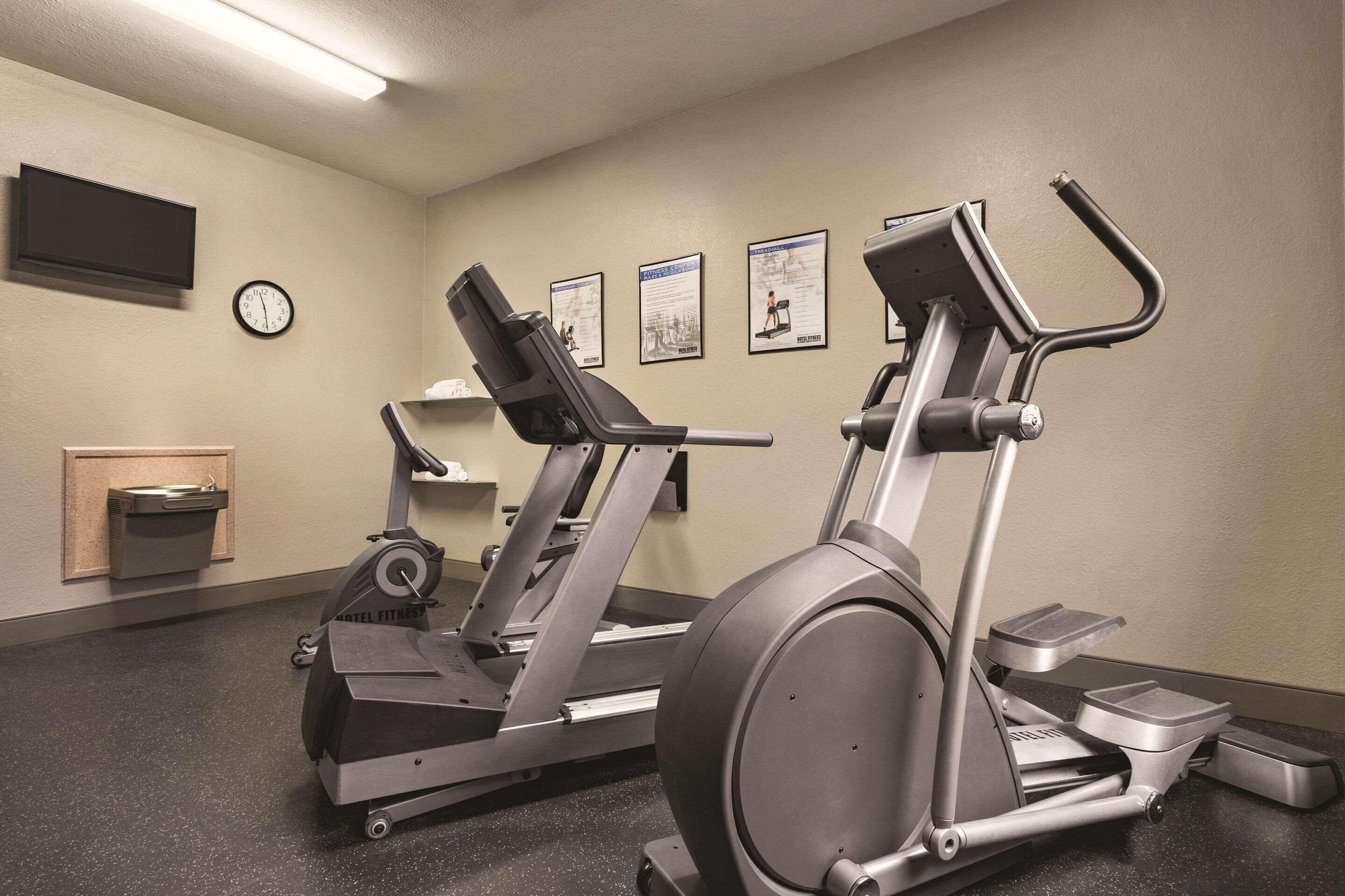 Un gimnasio con cintas de correr y elípticas, un reloj y carteles de ejercicios en la pared.