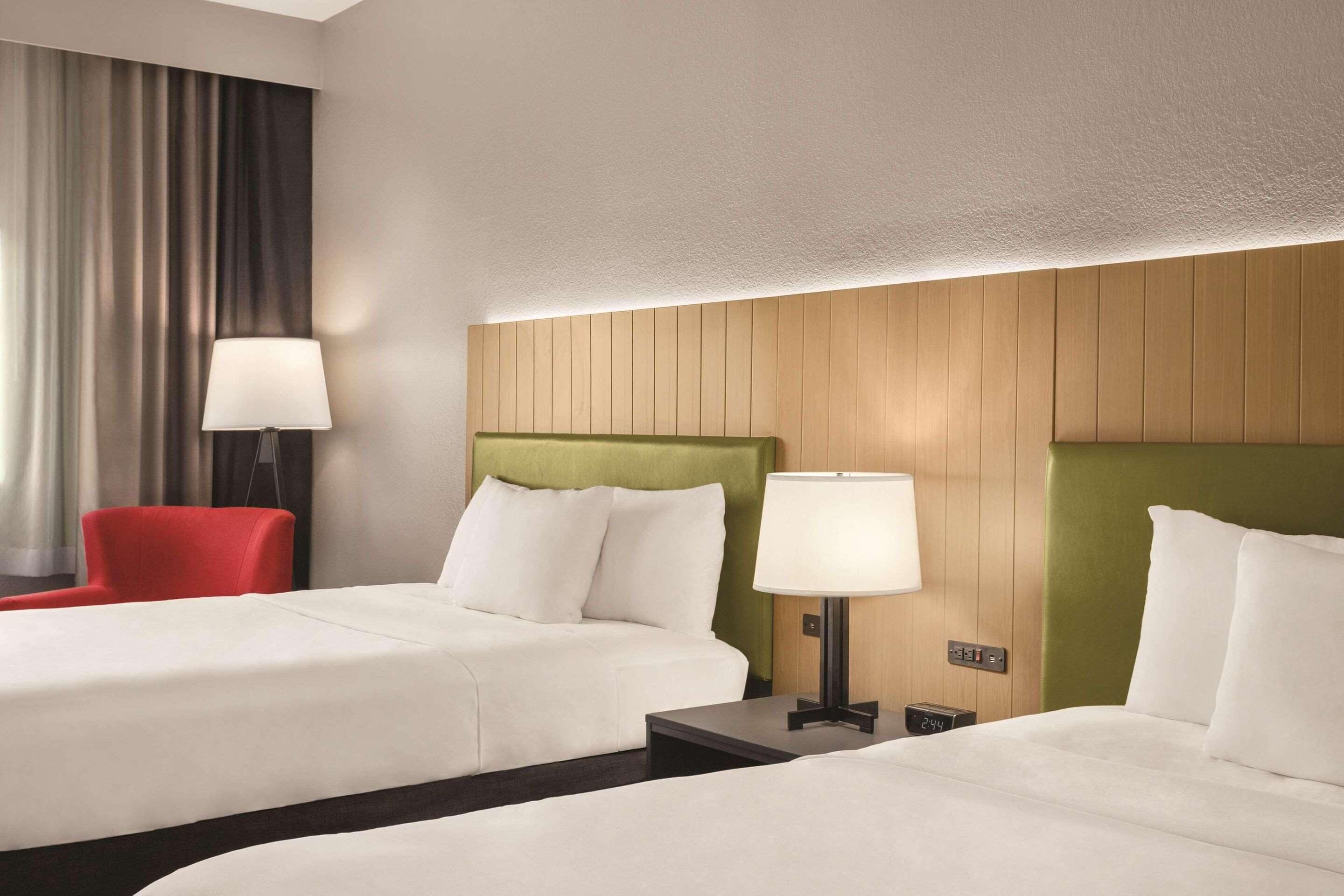 A hotel room with two beds, a red chair, and a lamp.