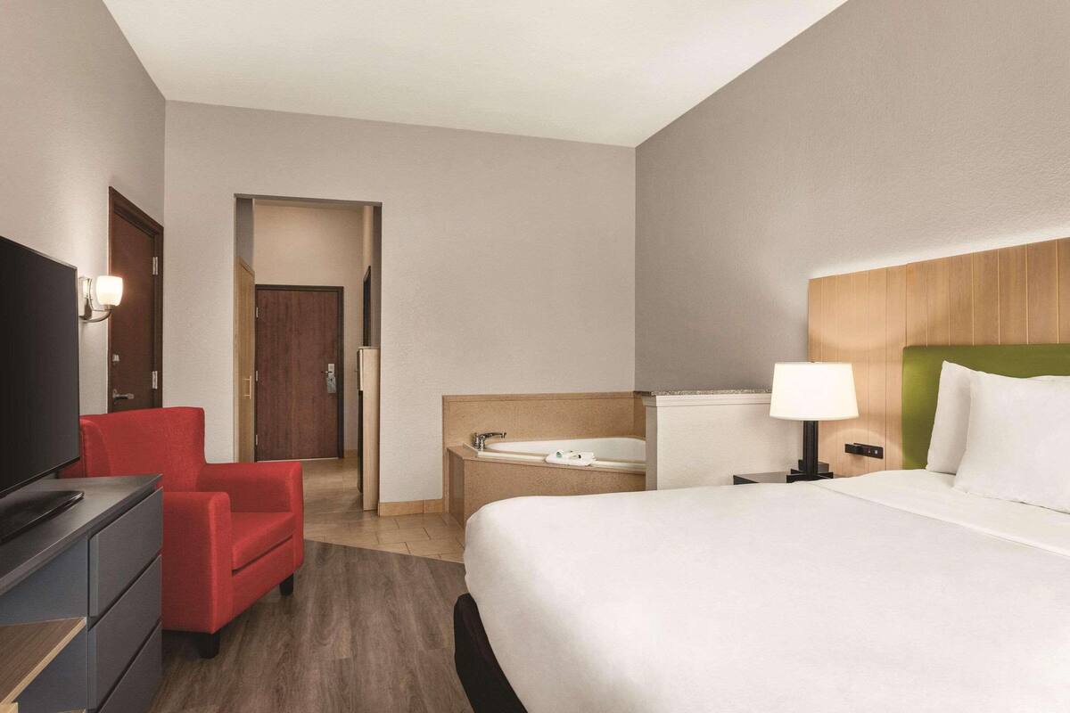 Habitación de hotel con cama, escritorio con televisor, una silla roja, baño con bañera y puerta que da a otra habitación.