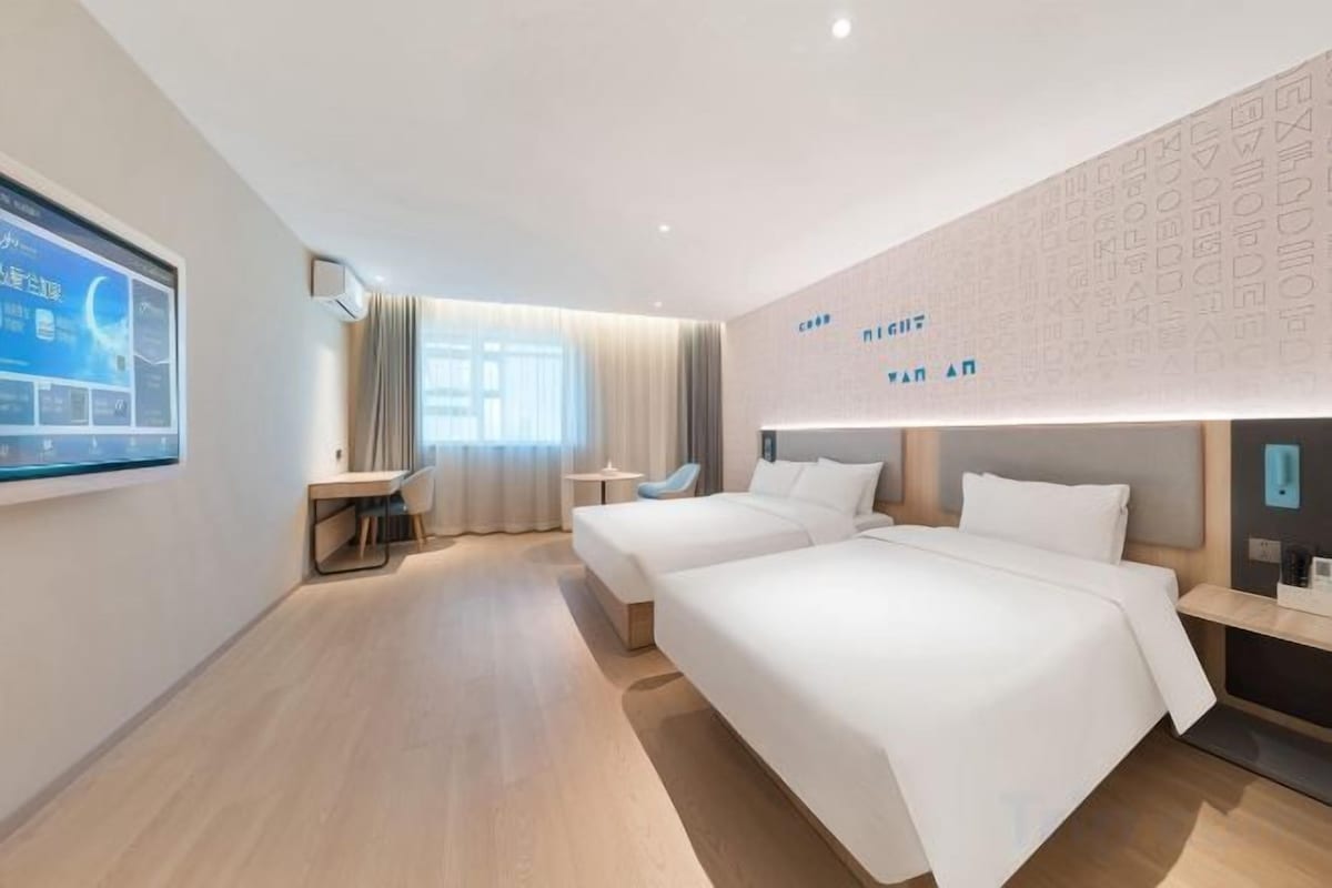 A hotel room with a desk, two beds, a large window, and a wall-mounted TV.