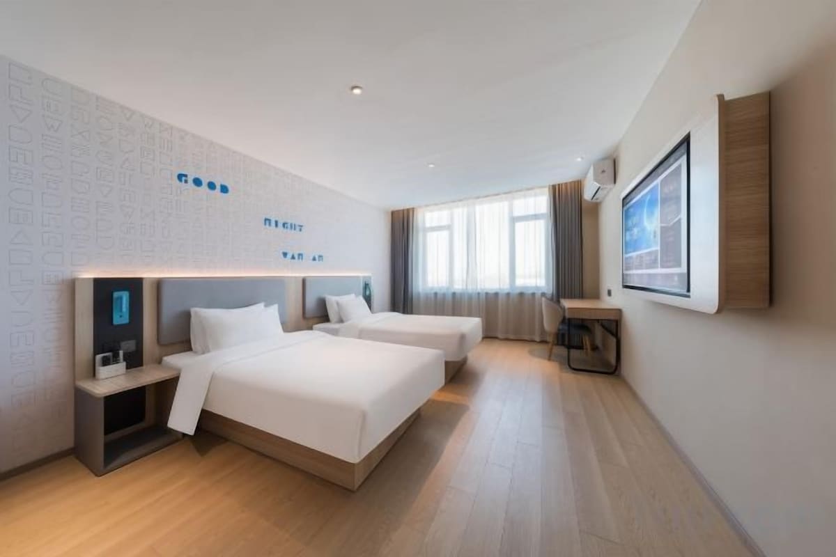 A hotel room with two beds, a bedside table, a wall-mounted TV, and a large window.