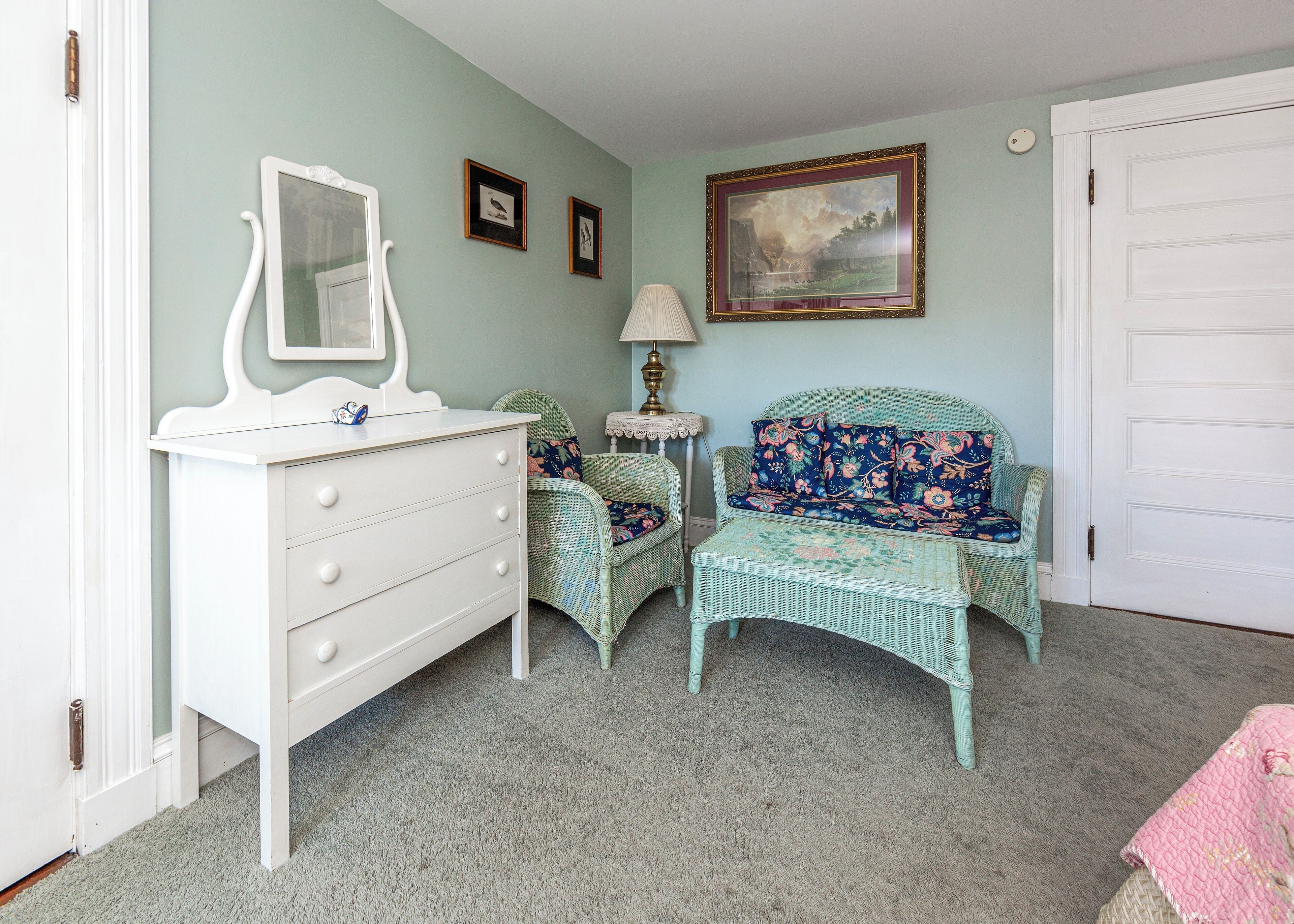 Een kamer met een witte dressoir, een rieten stoel met bloemenstoffering, een klein tafeltje met een lamp, en een lijst met een schilderij aan de muur.