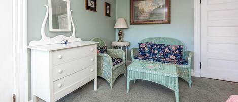 Een kamer met een witte dressoir, een rieten stoel met bloemenstoffering, een klein tafeltje met een lamp, en een lijst met een schilderij aan de muur.