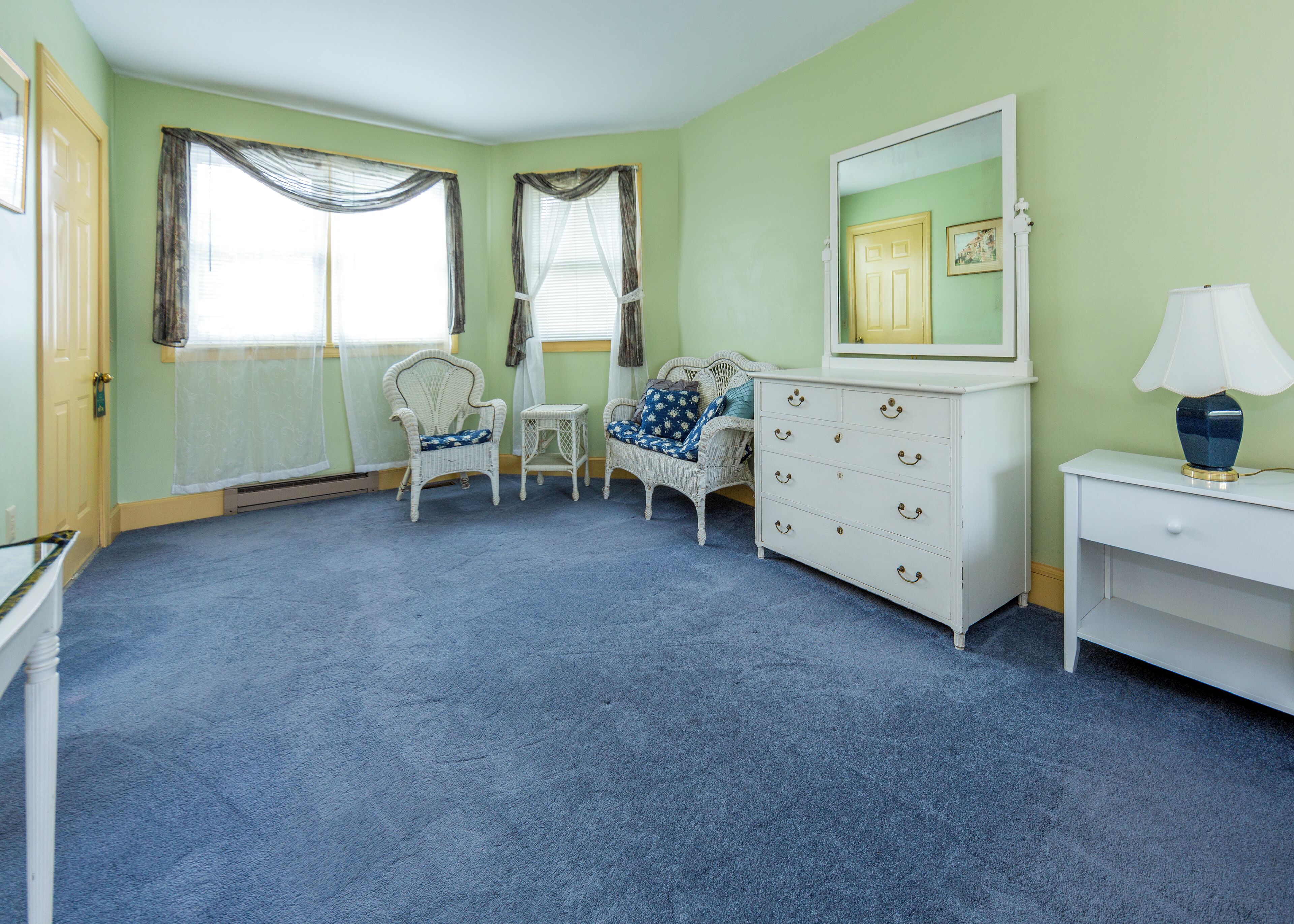 Une pièce avec un tapis bleu, une commode blanche à tiroirs, une chaise blanche et une petite table avec une lampe.
