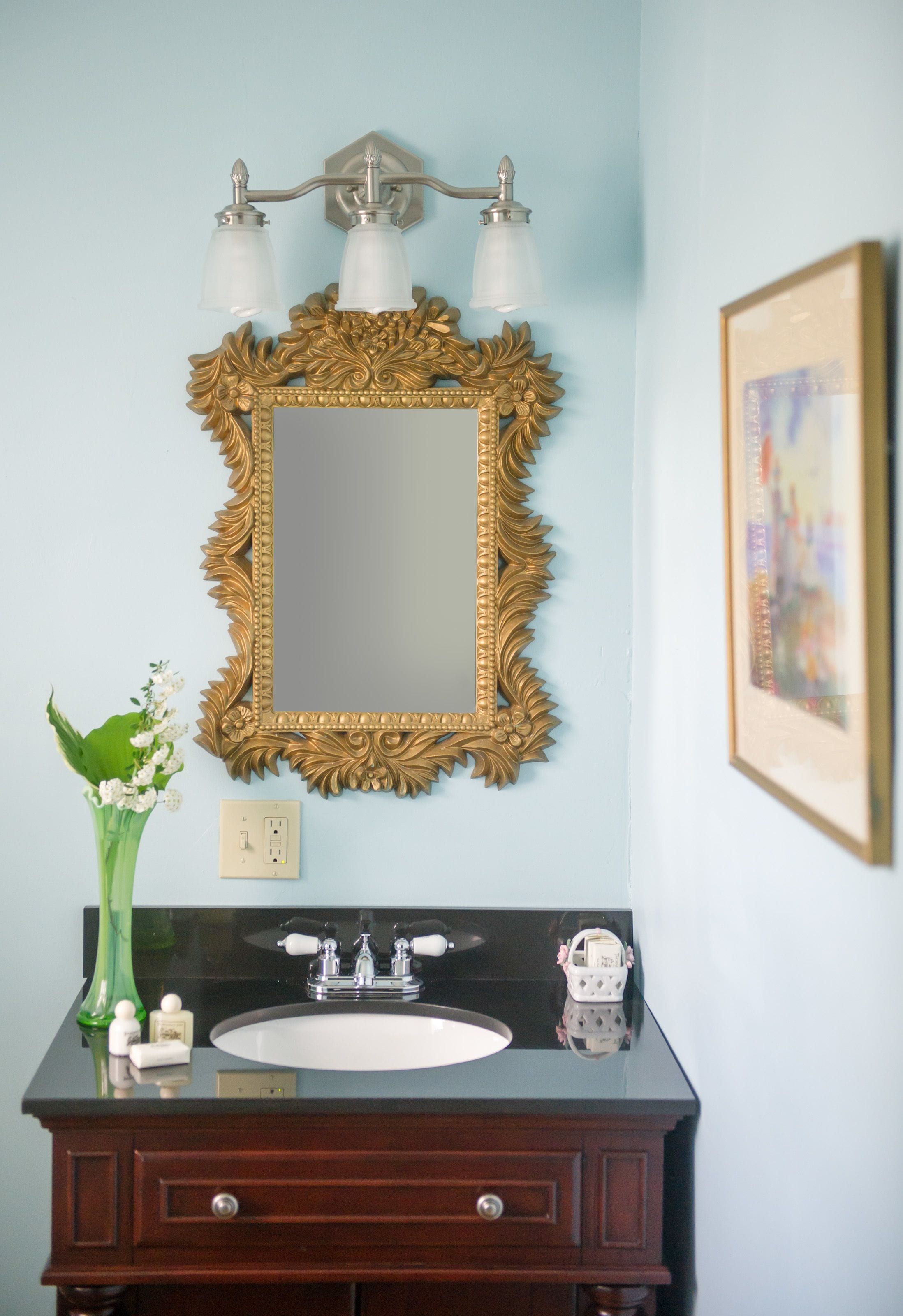 Un baño con un espejo de marco dorado, un lavabo negro y un florero verde con flores.