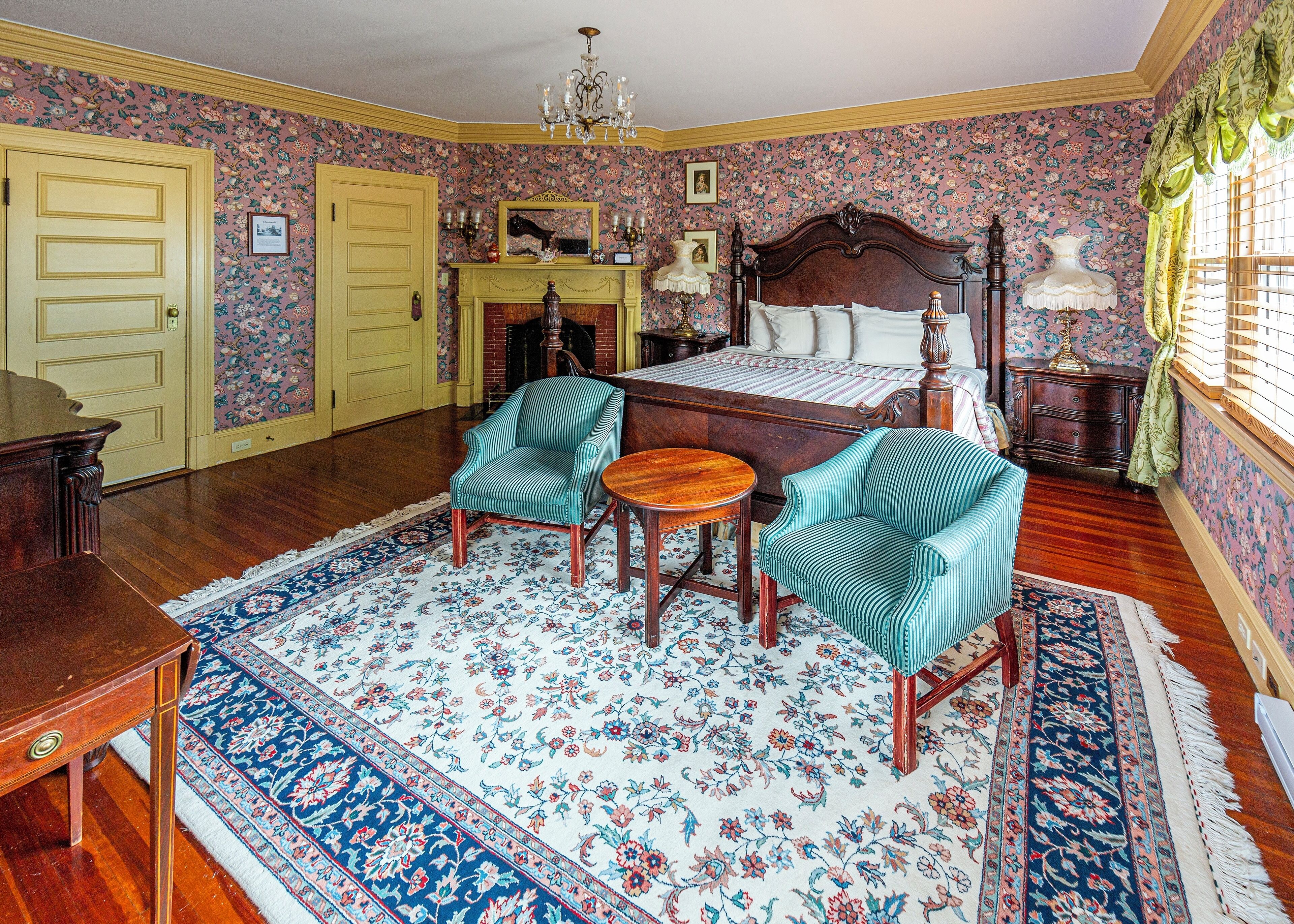 A room with a bed, two chairs, a table, a fireplace, and a chandelier.