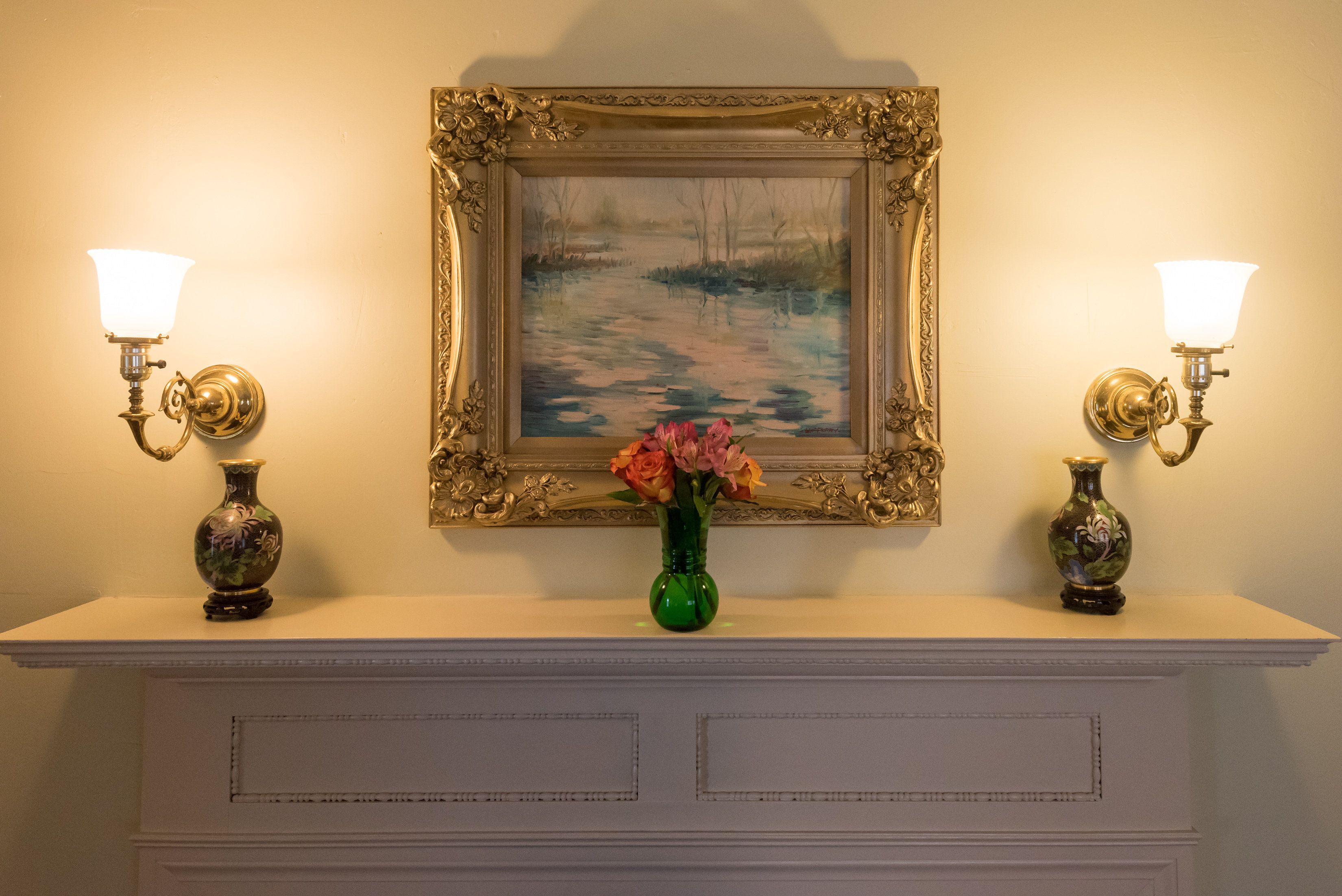 A framed painting on a wall, two wall sconces, and a vase with flowers on a mantel.