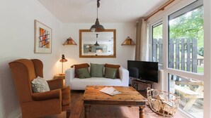 Sala de estar com sofá branco, poltrona marrom, mesa de centro de madeira e uma TV em um suporte.