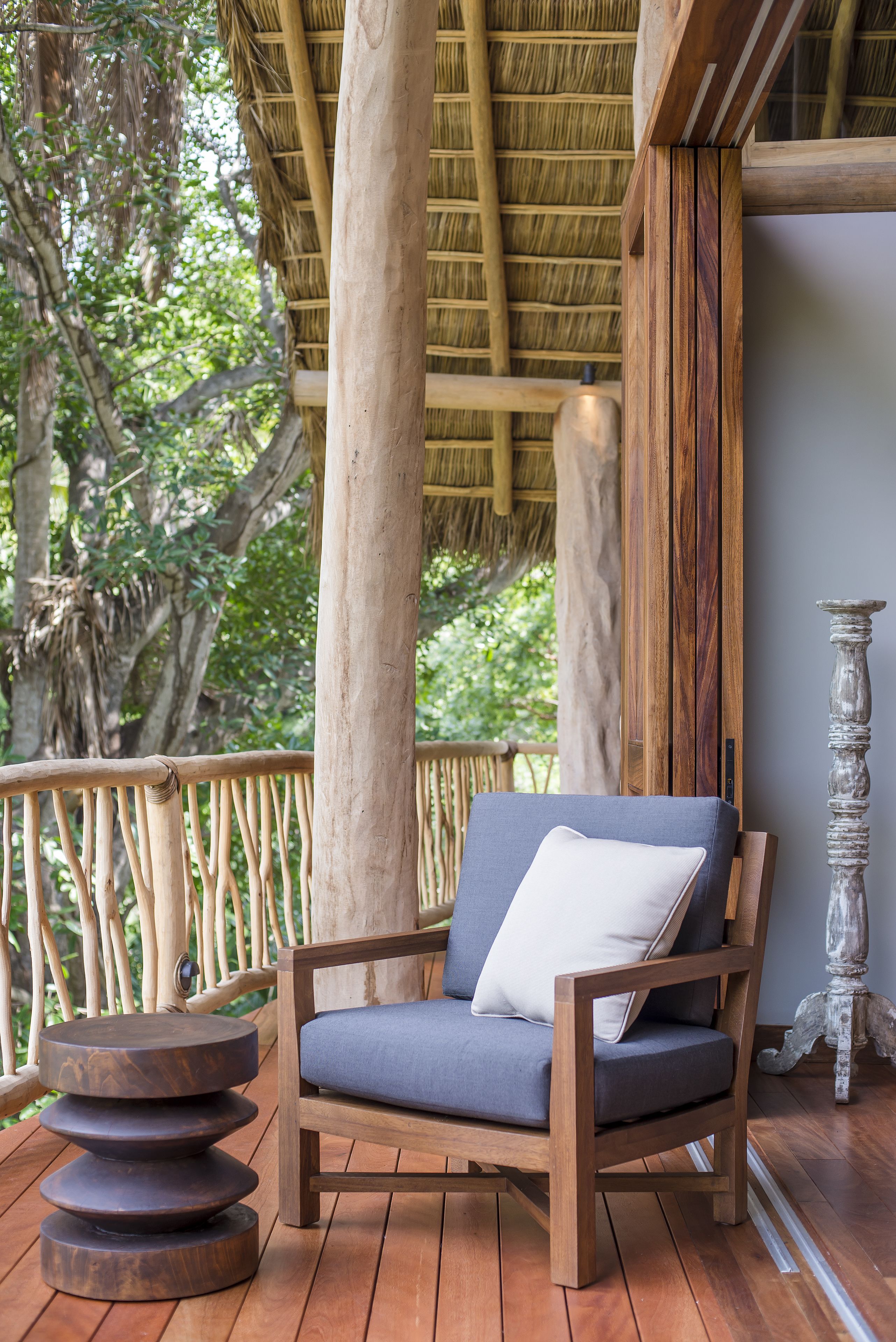 A wooden chair with a cushion on a wooden deck, surrounded by a thatched roof structure and wooden pillars.