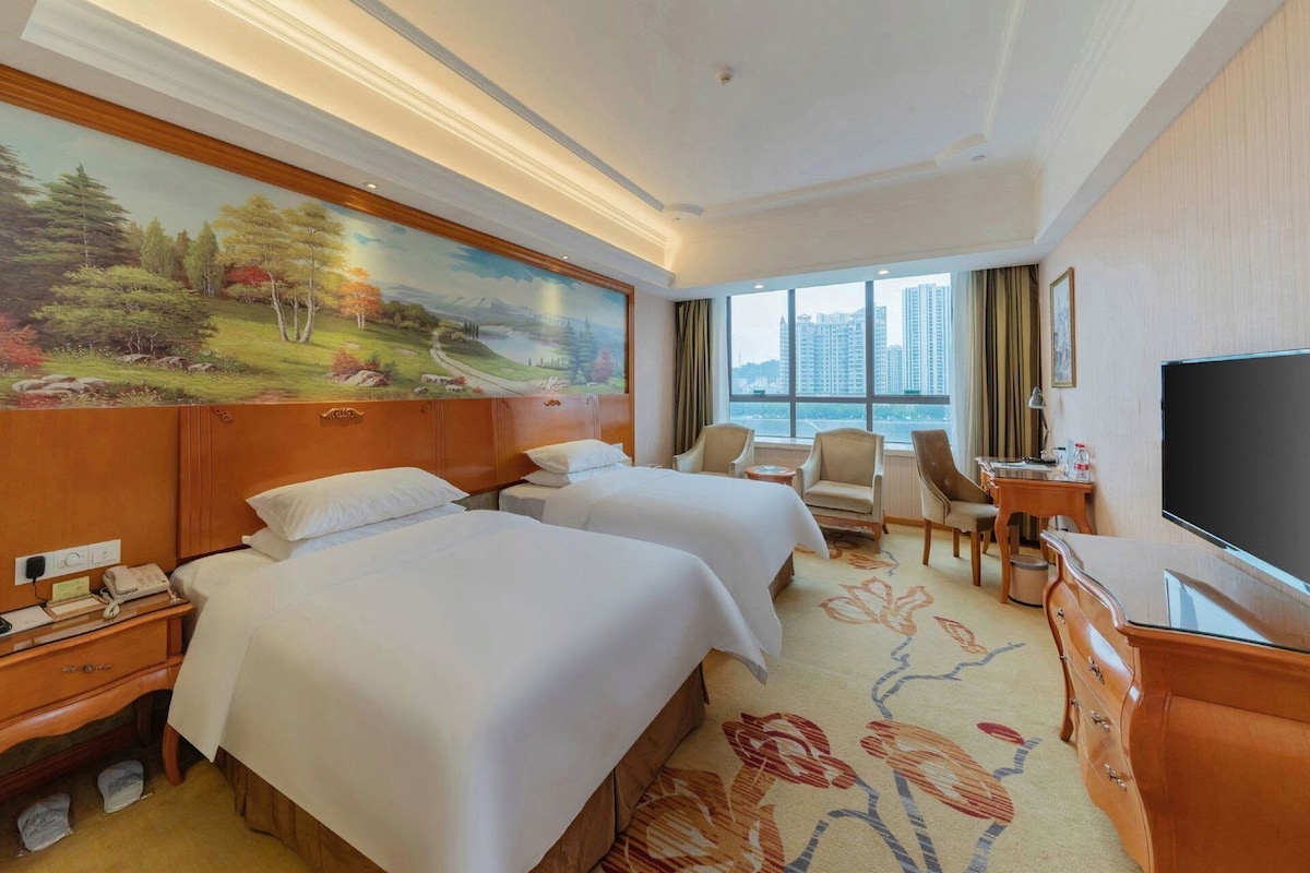 A hotel room with two beds, a desk, a chair, a television, and a large window with a city view.
