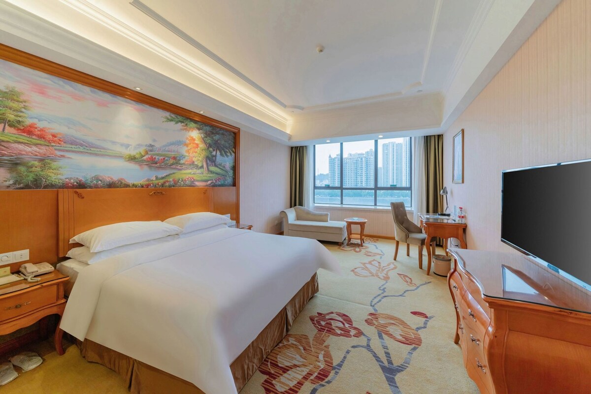 A hotel room with a large bed, a desk, a chair, a television, and a painting on the wall.
