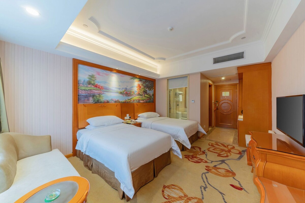 A hotel room with two beds, a sofa, a TV, and a painting on the wall.