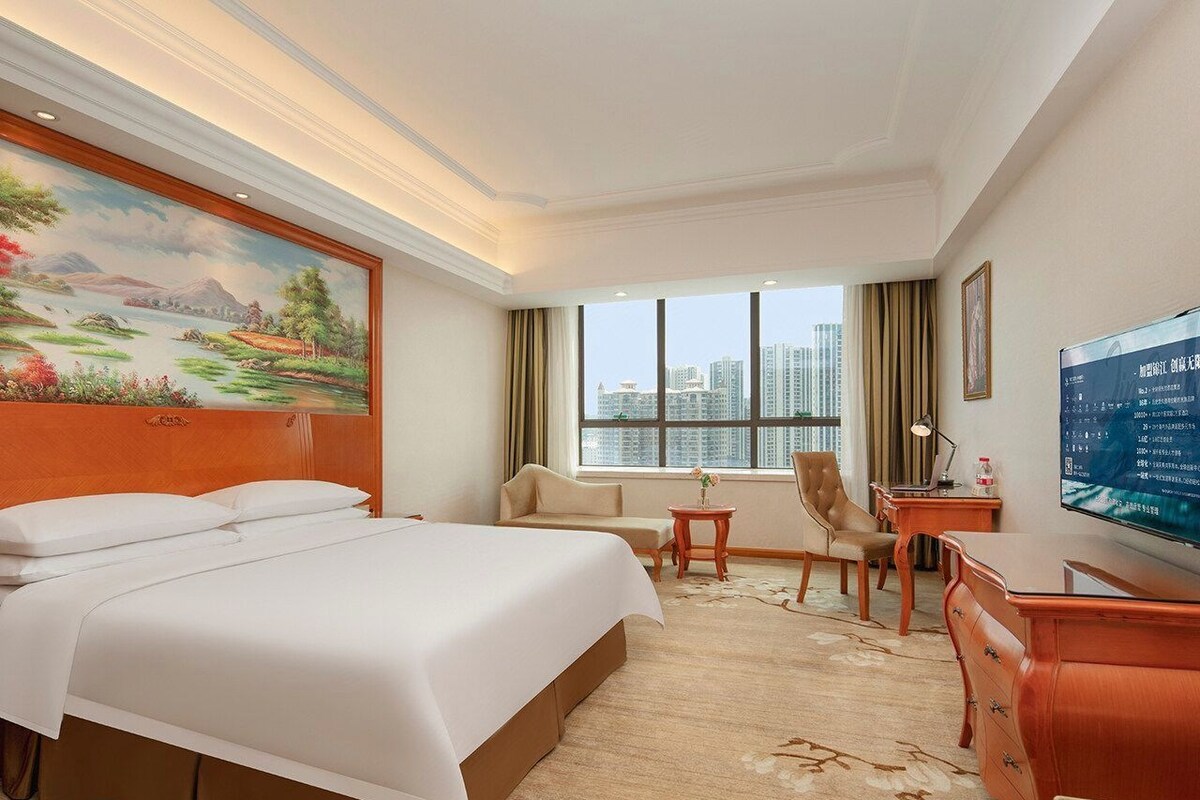 A hotel room with a large bed, a desk, a chair, a TV, and a window with a city view.