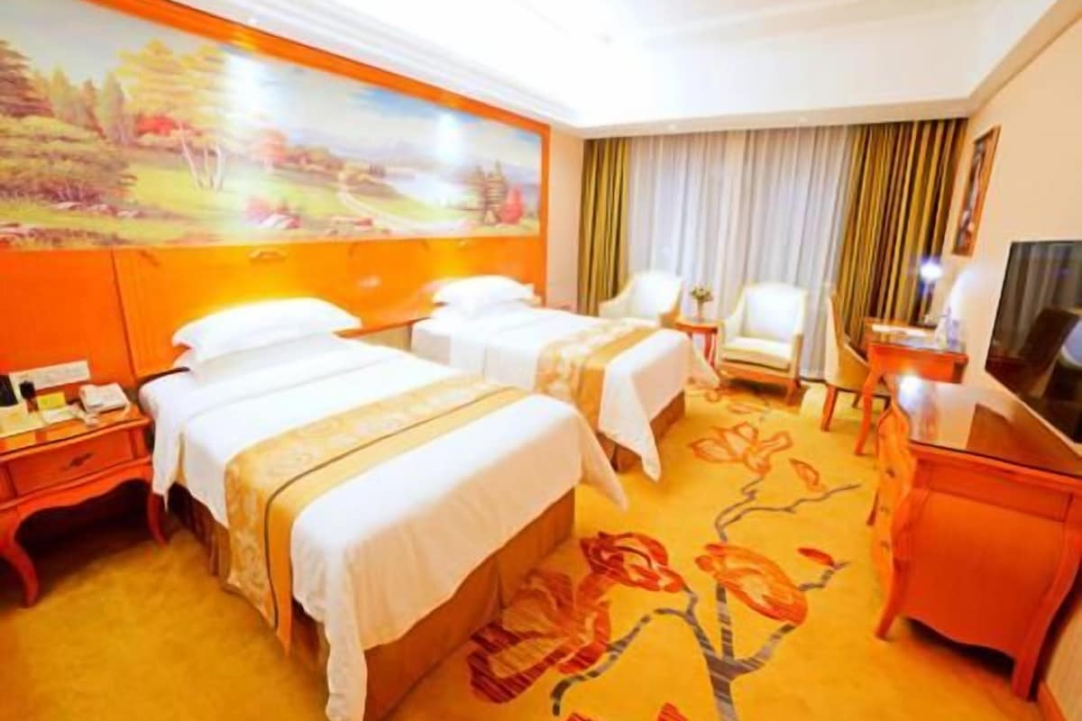 A hotel room with two beds, a desk, a chair, a TV, and a painting on the wall.