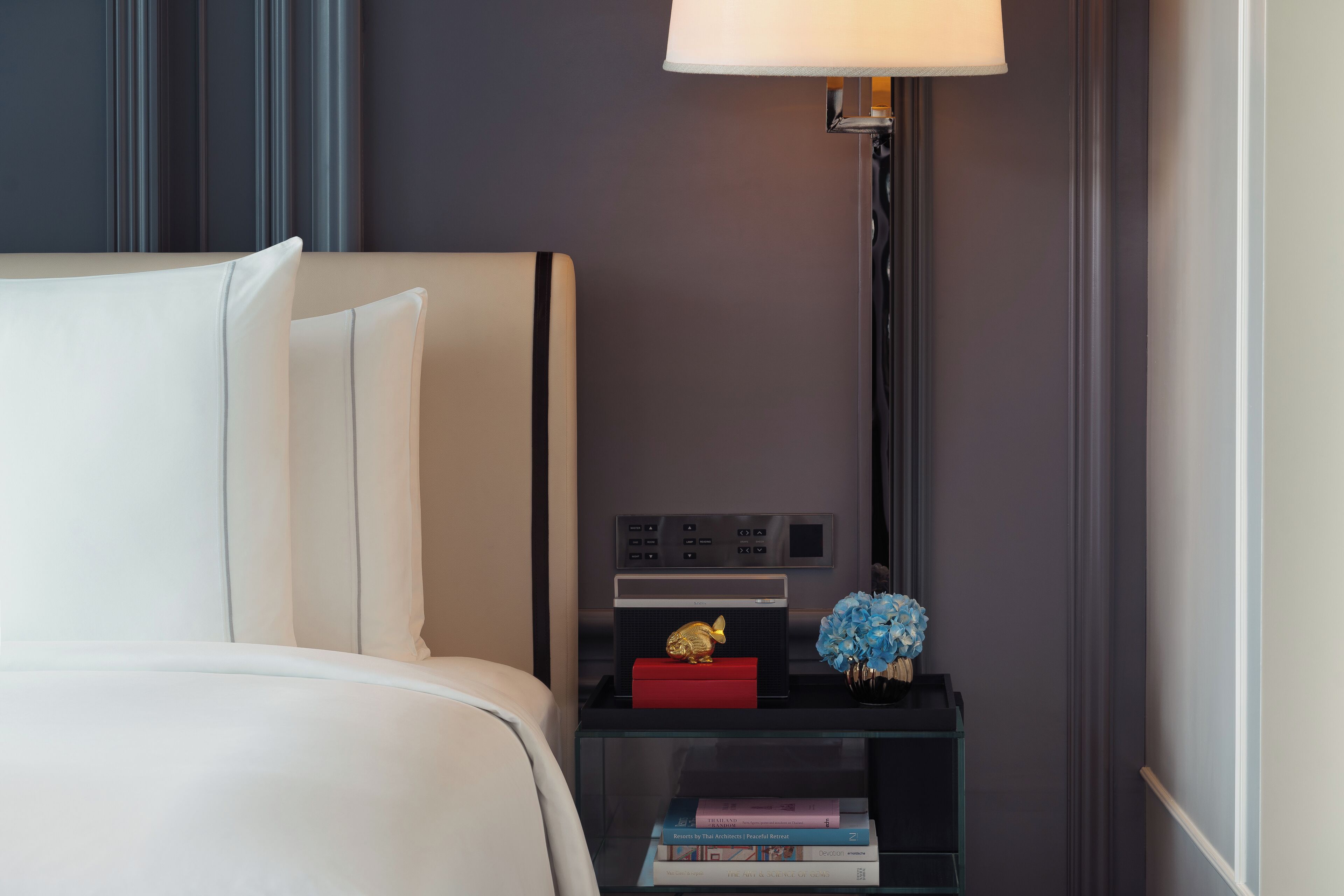 A neatly made bed with white linens, a bedside table with a lamp, a small red box, and a few books.