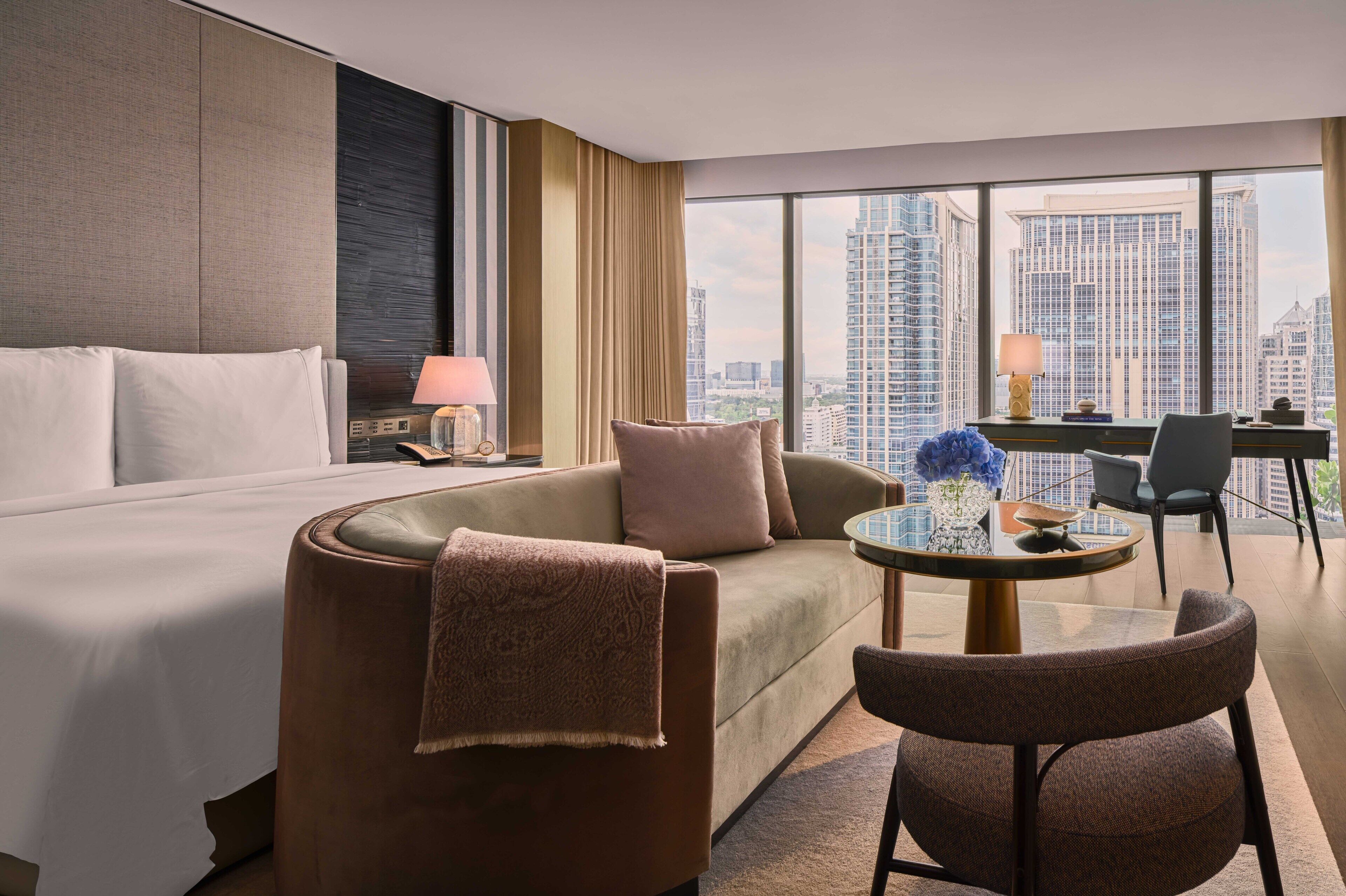 Ein modernes Hotelzimmer mit einem großen Bett, einer Couch, einem runden Tisch und einem Schreibtisch mit Lampe. Das Zimmer verfügt über große Fenster mit Blick auf die Stadt.