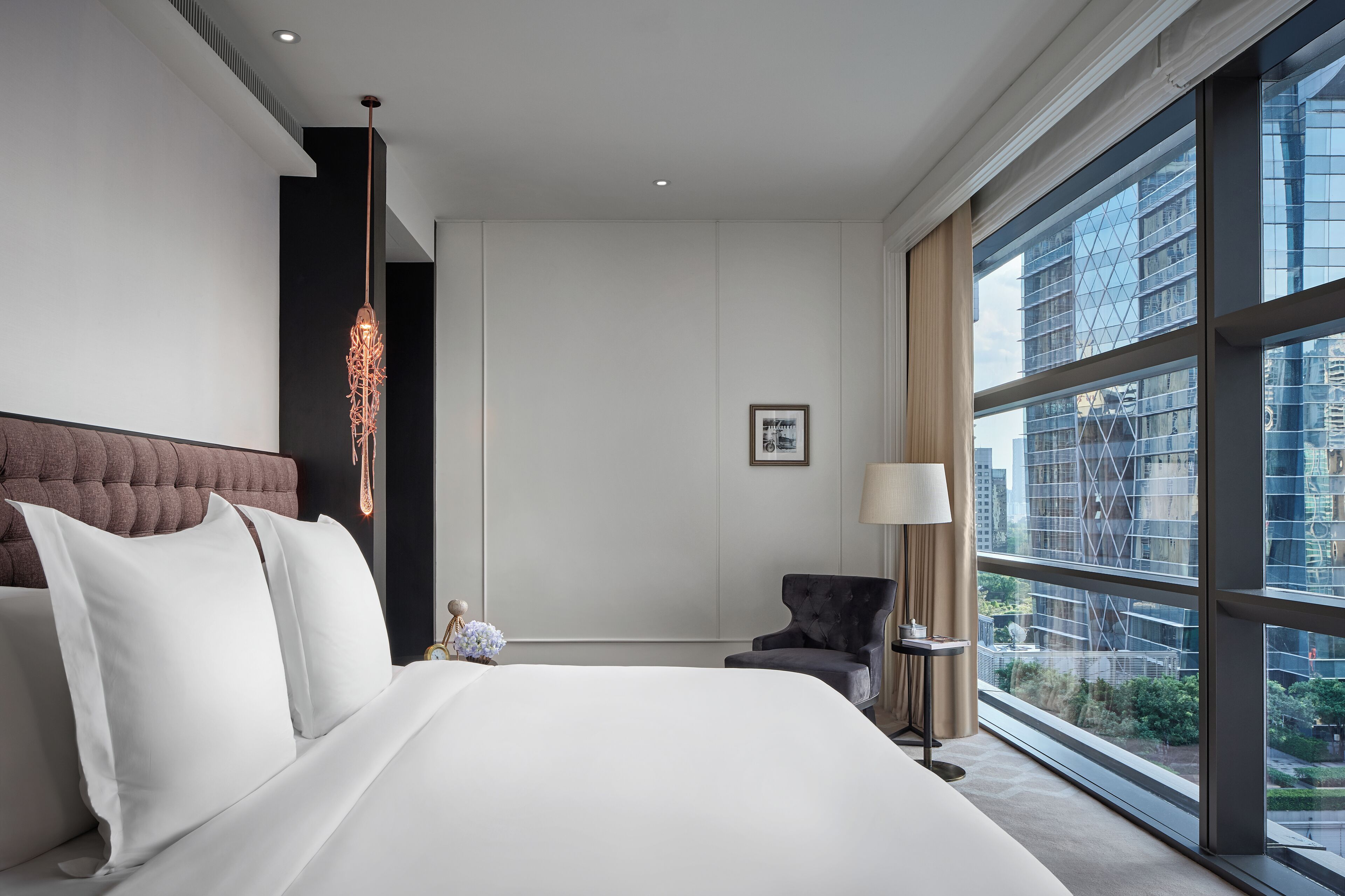 A hotel room with a large bed, a modern chandelier, a view of a cityscape through large windows, and a small armchair.