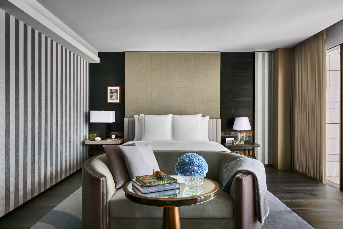 Una habitación de hotel moderna con una cama grande, una mesita de centro redonda con libros y un jarrón, una mesita de noche con una lámpara, y vistas de la ciudad a través de amplias ventanas.