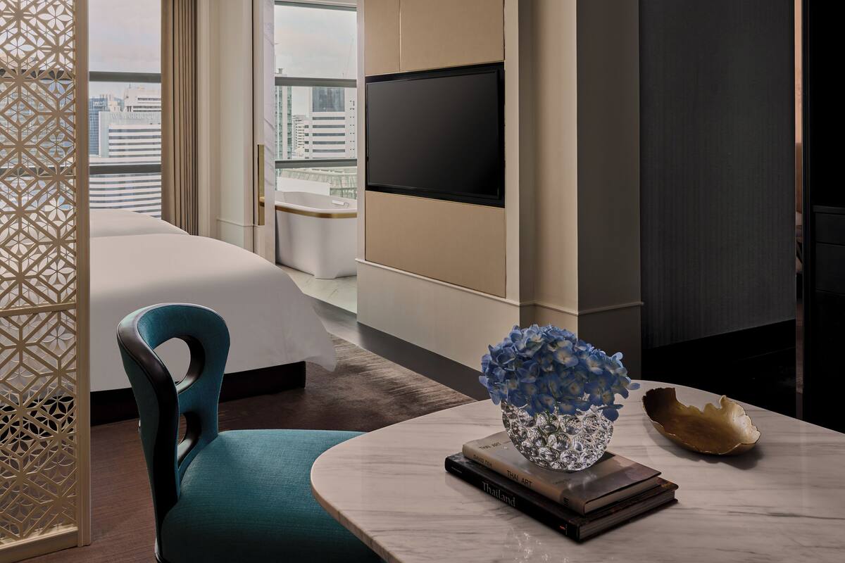 Una habitación de hotel moderna con cama, televisor montado en la pared, una mesa de comedor con un libro y objetos decorativos, y vista del paisaje urbano a través de amplias ventanas.