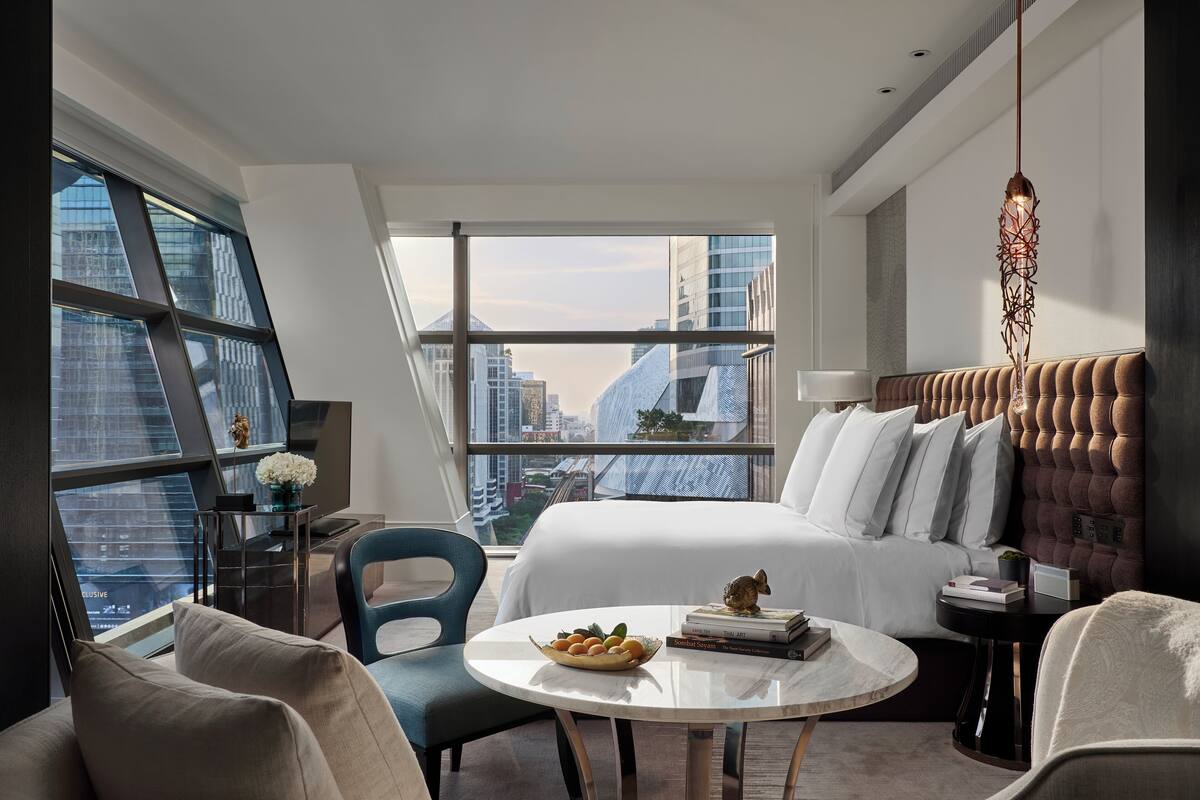 Una habitación de hotel moderna con una cama grande, una mesa de comedor redonda y vistas a un paisaje urbano a través de ventanales de piso a techo.
